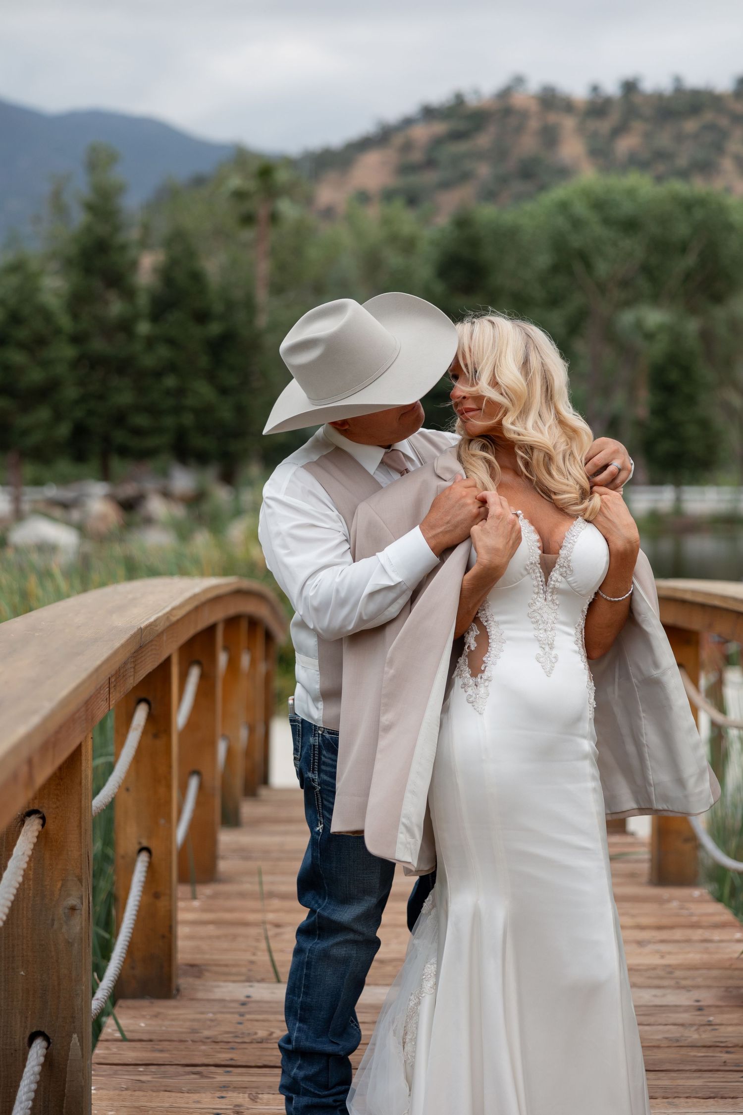 couple in wedding attire, embracing back to front and looking at each other on a wooden bridge with green hills in back.