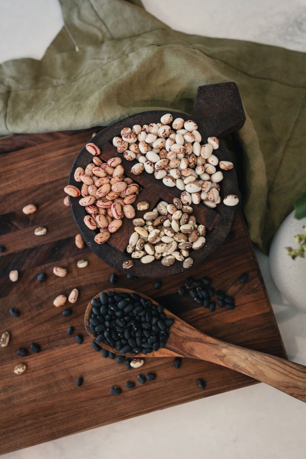 Various dried beans and legumes displayed on wooden cutting board with olive green fabric.