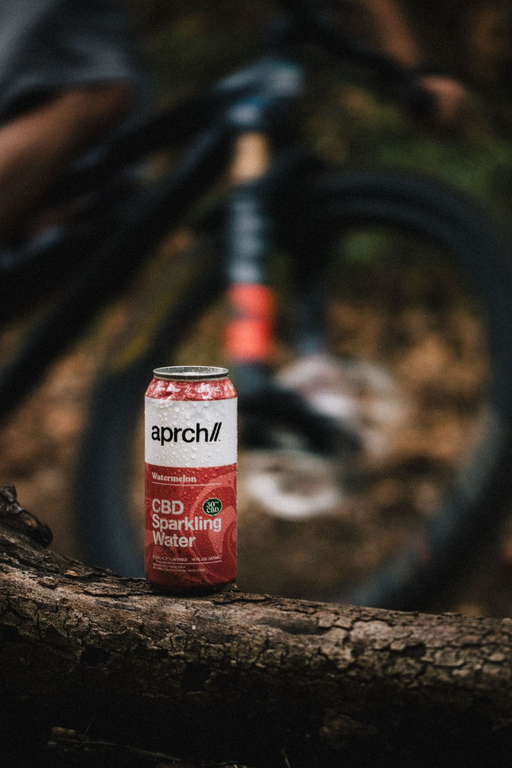 Aprch CBD beverage can placed on a log with a mountain bike blurred in the background.