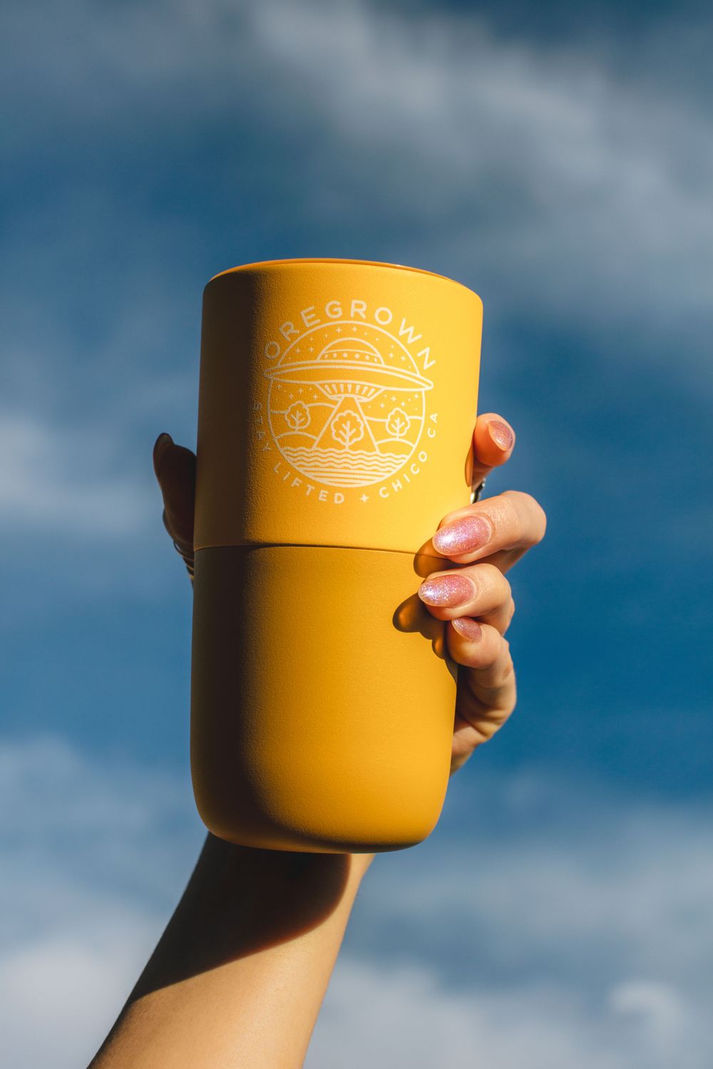Yellow reusable coffee cup held against blue sky with clouds.