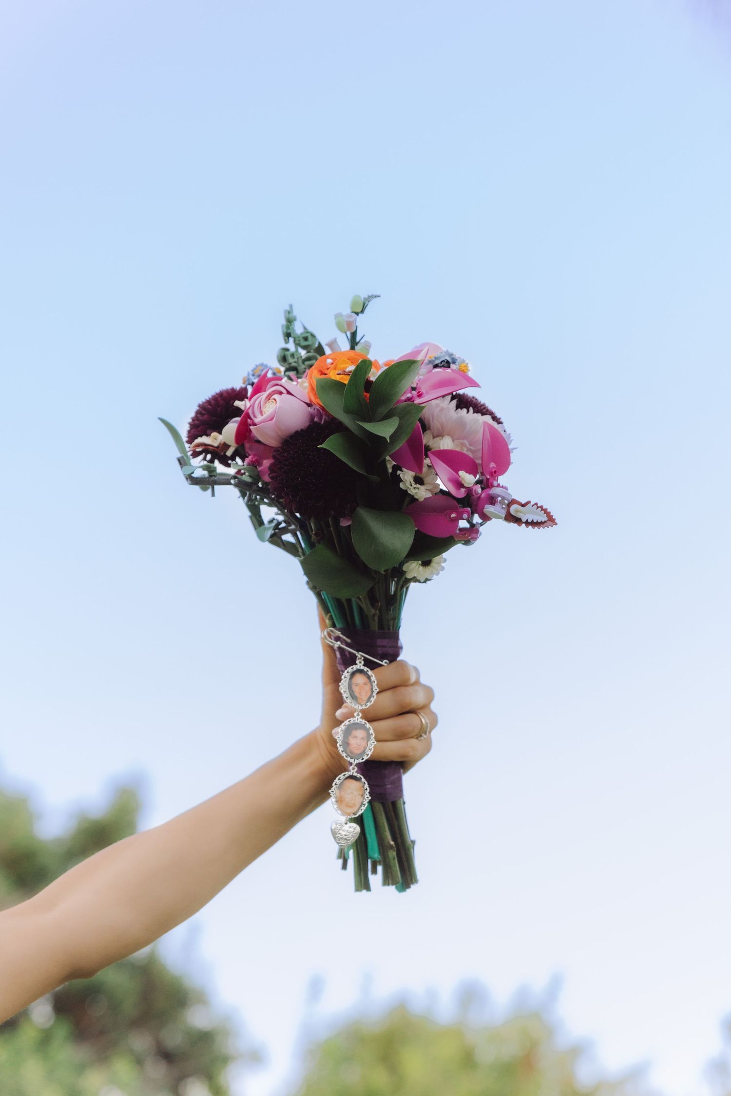 Hand raises a vibrant pink and purple bridal bouquet with photo charm against blue sky.