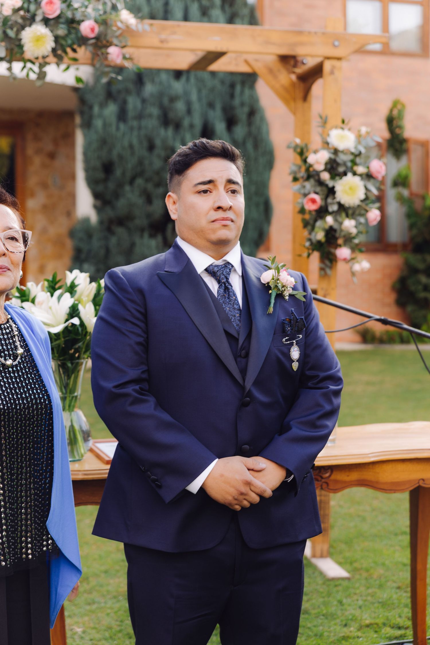 Groom in navy suit with boutonniere stands at outdoor wedding altar awaiting bride.