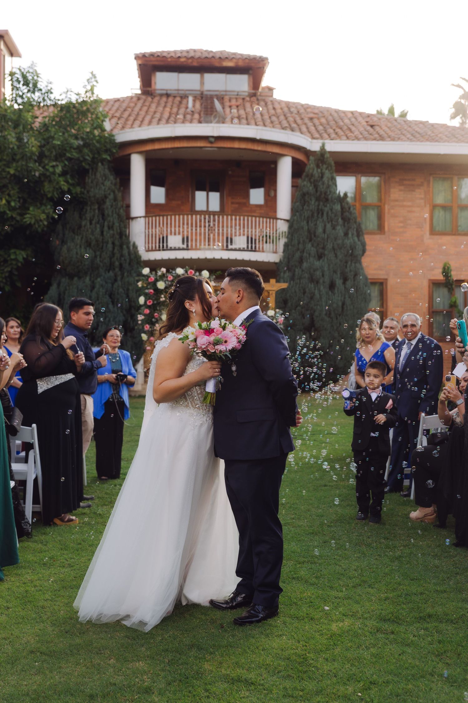 Newlyweds share first kiss on lawn while guests celebrate with bubbles at outdoor wedding.
