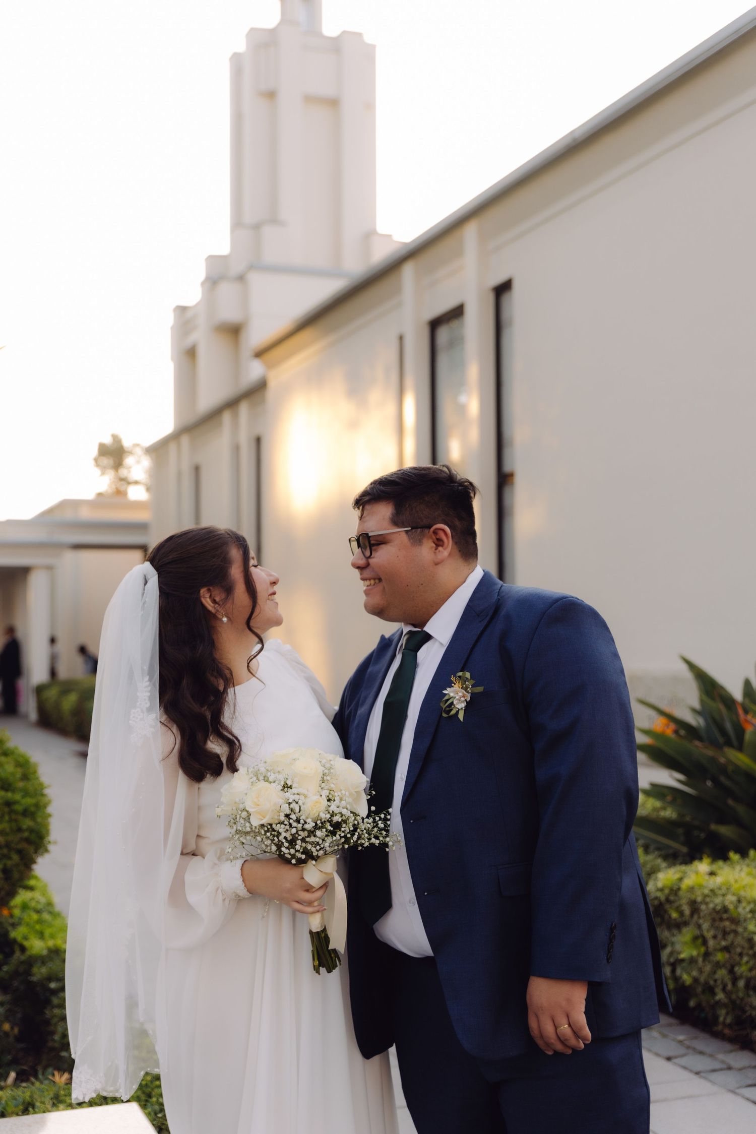 Bride in white gown and veil with bouquet stands beside groom in blue suit outside church.
