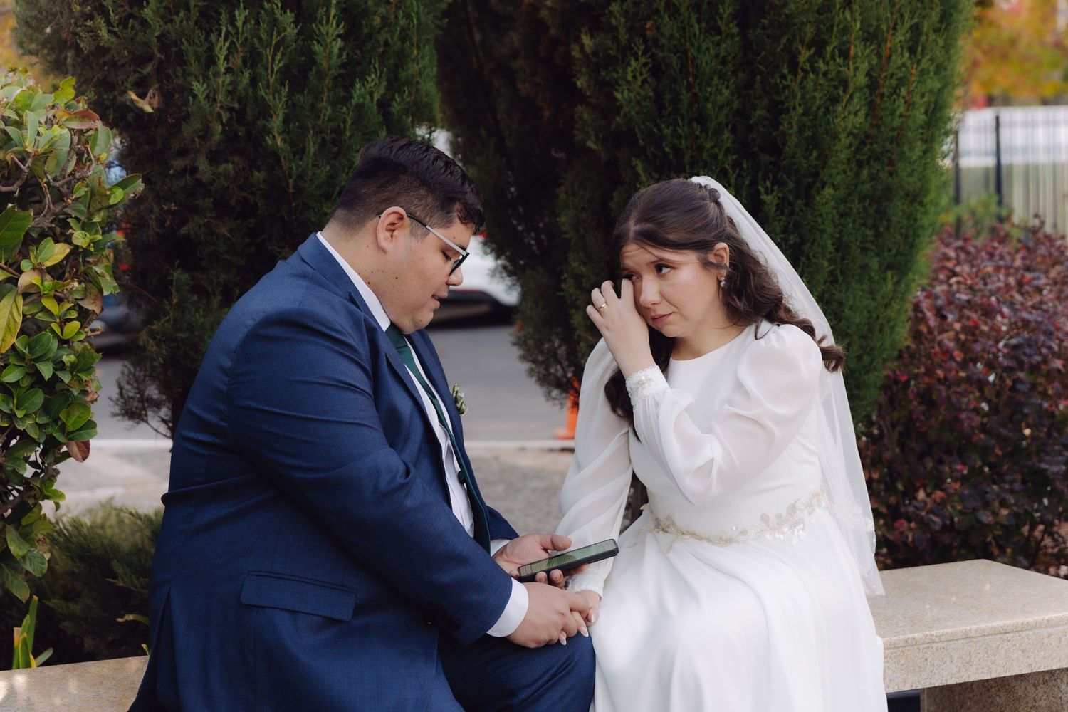 Emotional bride in white wedding dress wipes tears while groom holds her hands on bench.