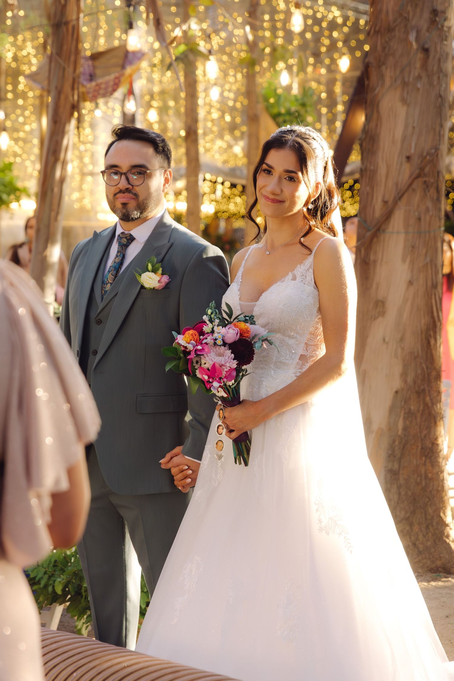 Bride in white lace gown and groom in gray suit hold hands during outdoor wedding ceremony.