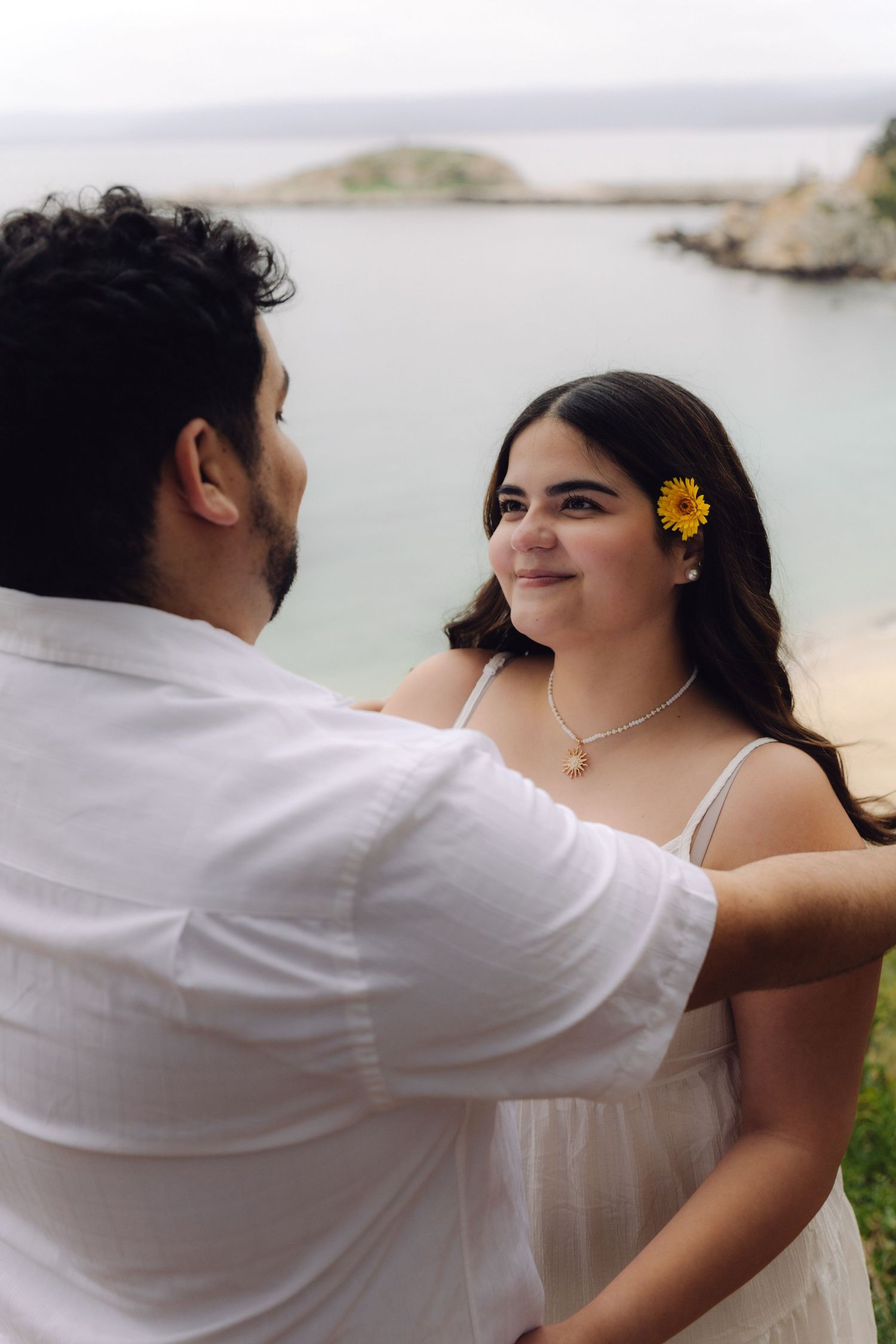 Couple holding hands near ocean, woman wearing sunflower in her hair.