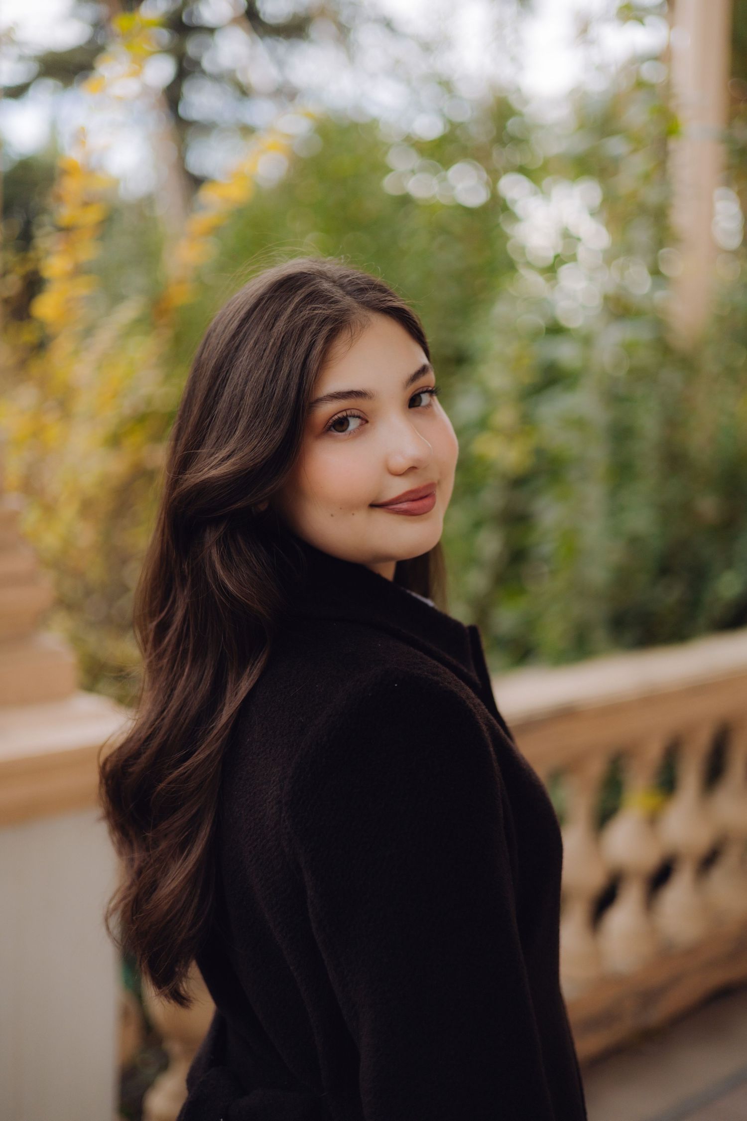 Young brunette woman in black coat smiling over shoulder in autumn garden setting.