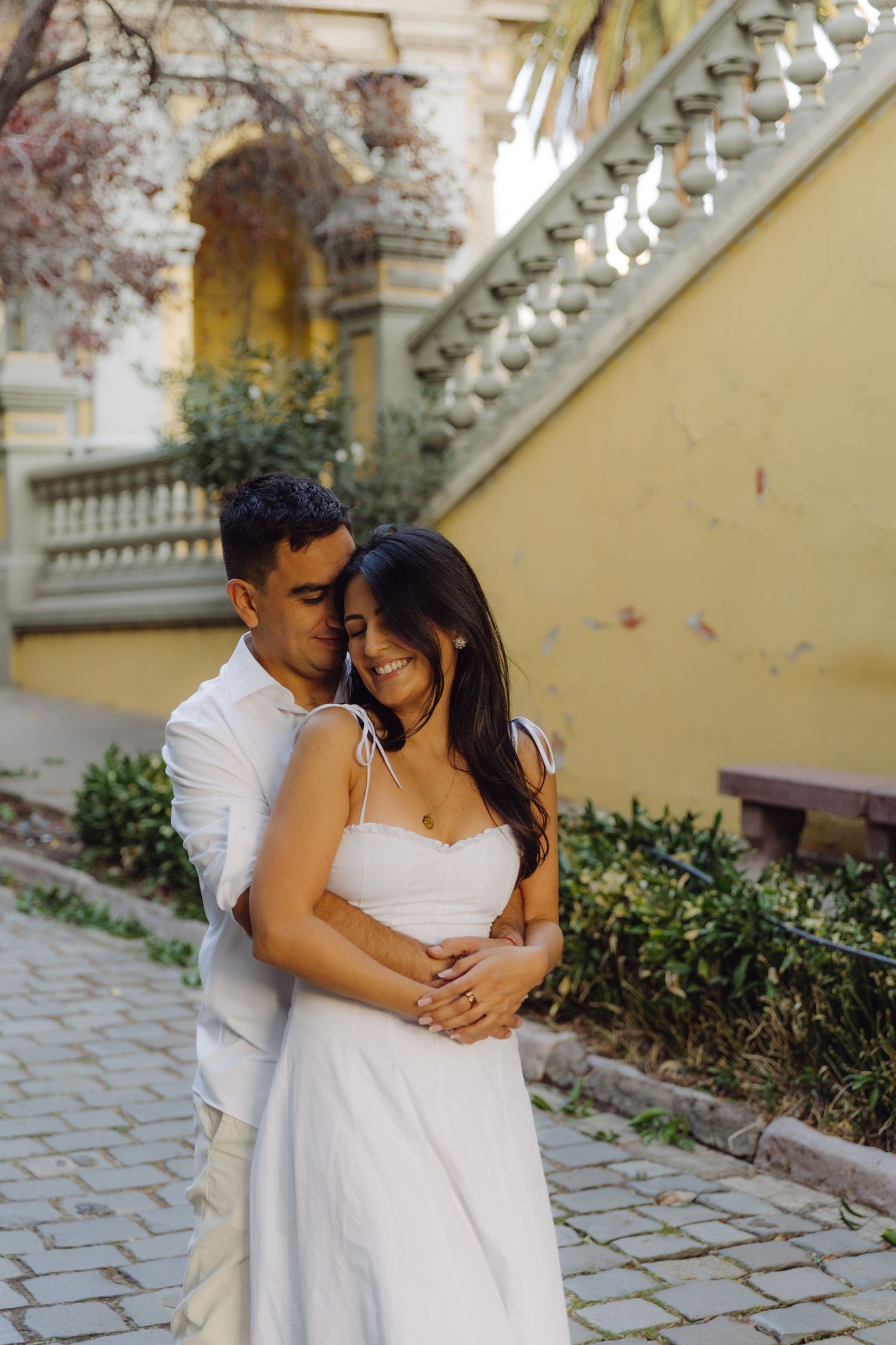 Couple embracing tenderly in front of a yellow historic building with ornate staircase and blooming trees.
