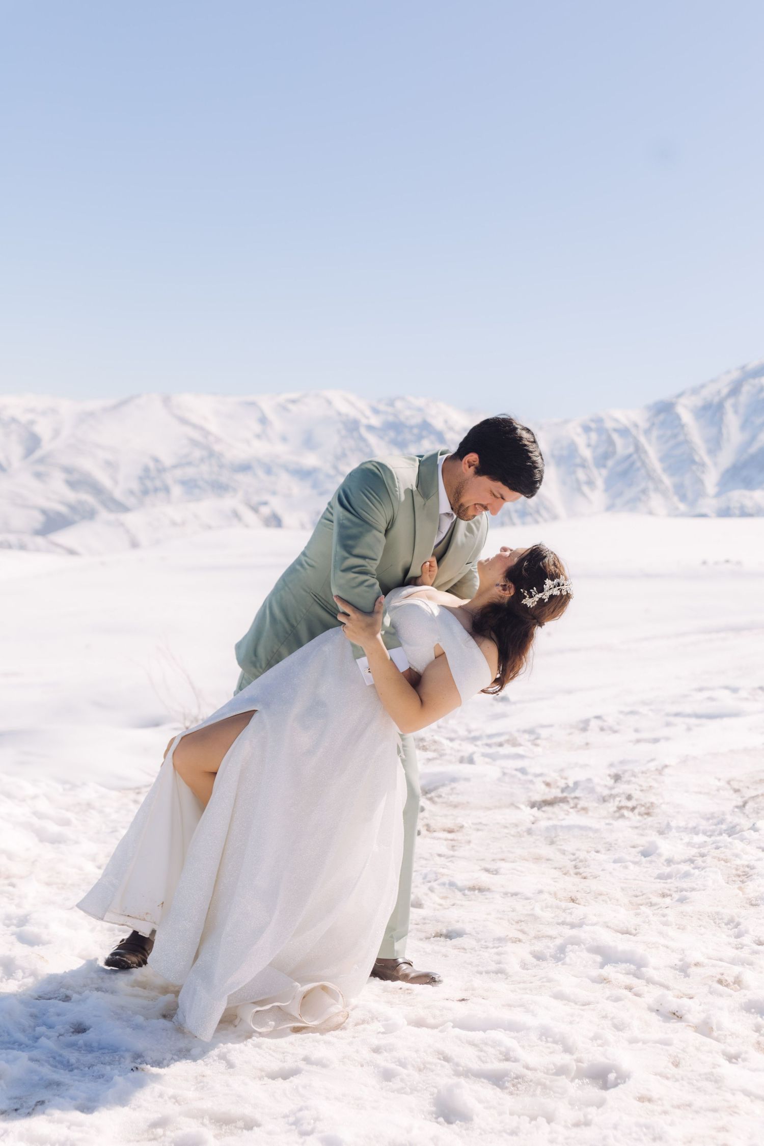 Bride and groom sharing a romantic dip kiss on a snowy mountain landscape in winter wedding attire.