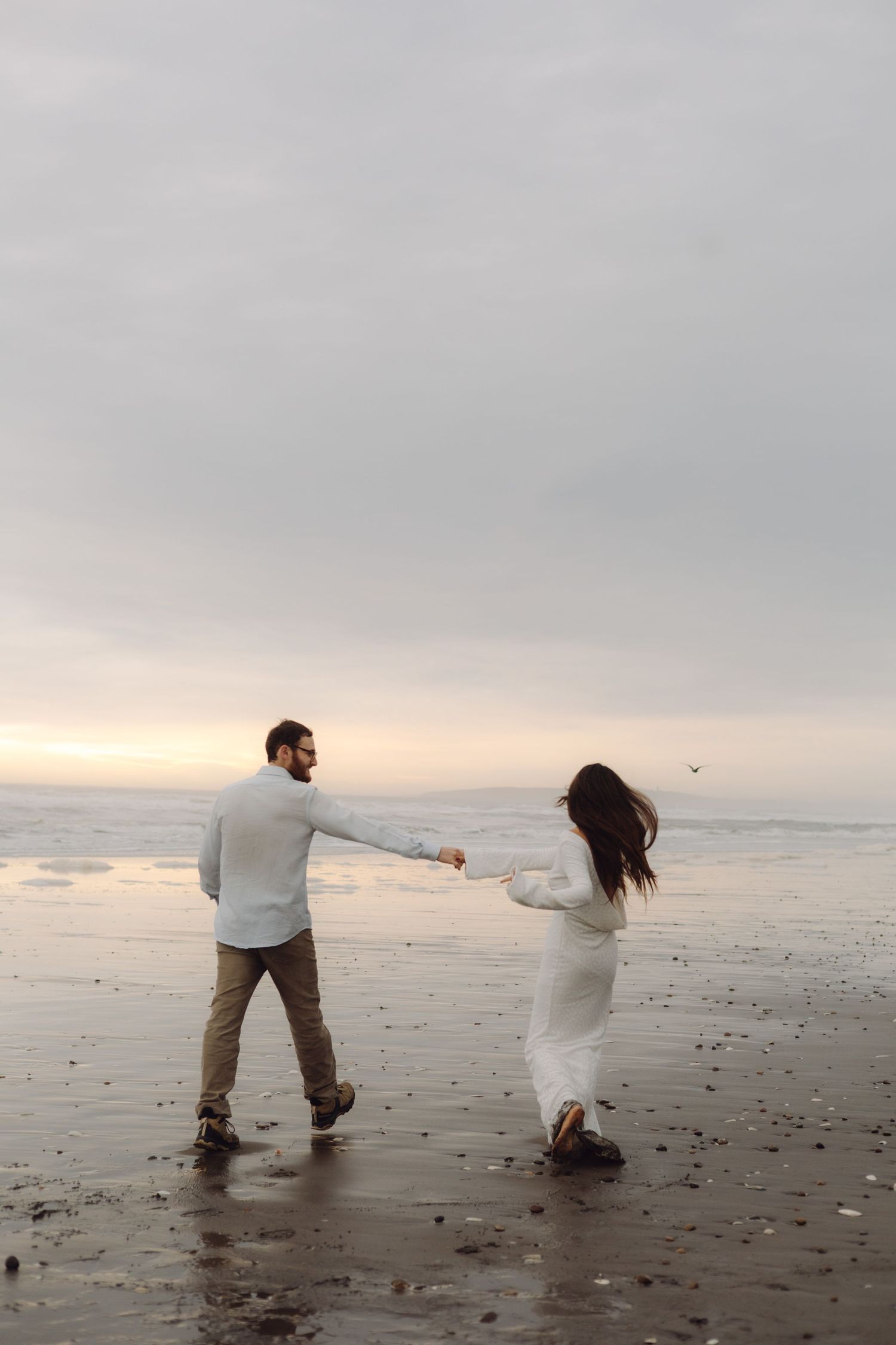Couple holding hands on a misty beach at sunset, woman in white dress being led toward the ocean.