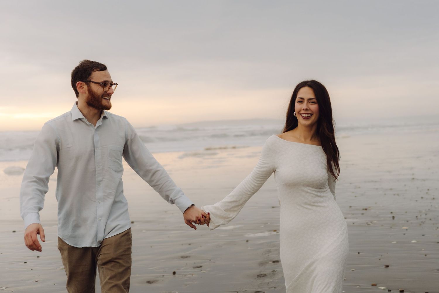 Happy couple holds hands walking on beach at sunset, woman in white dress, man in light shirt.
