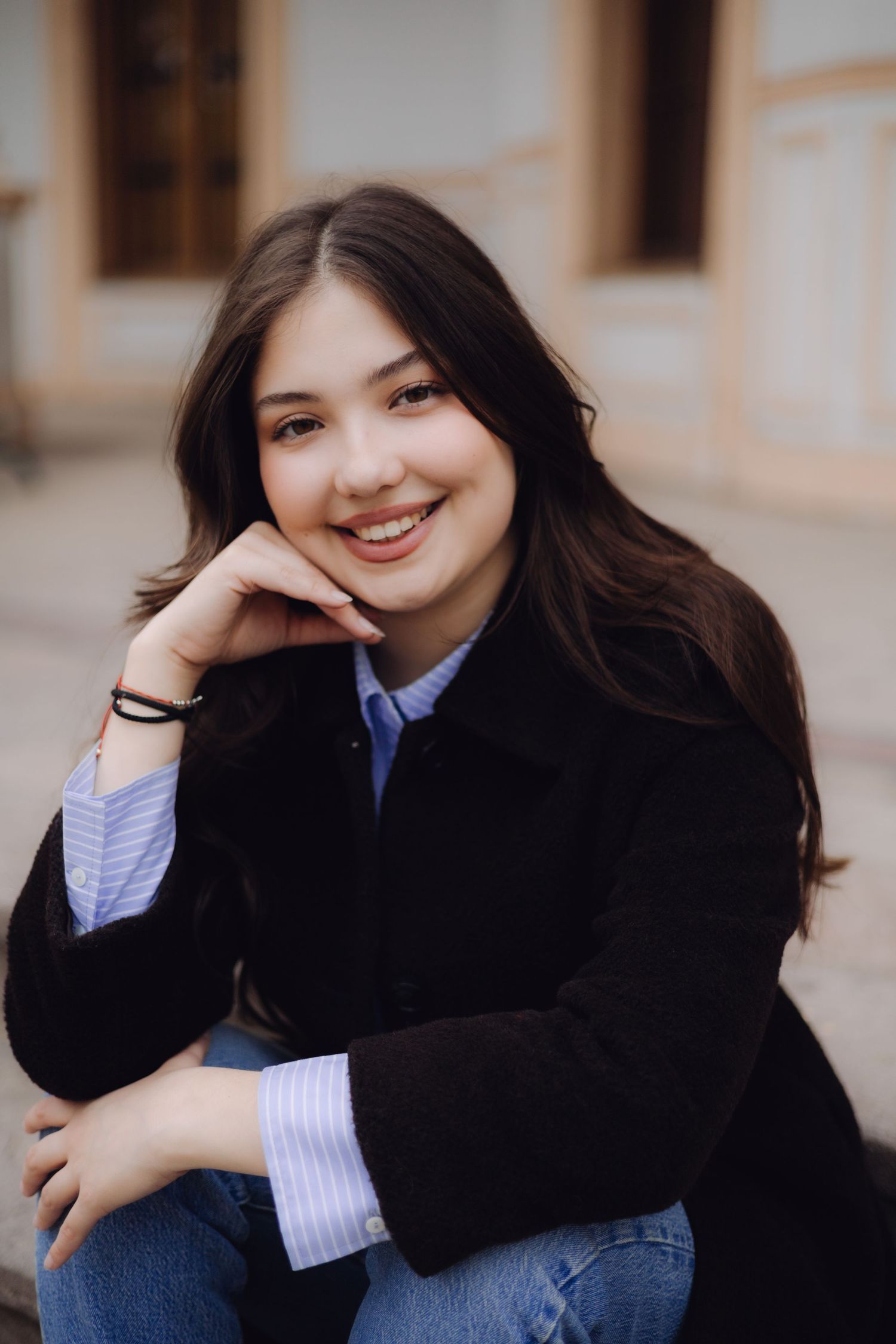 Smiling young brunette woman in black blazer and blue shirt sitting outdoors on steps.