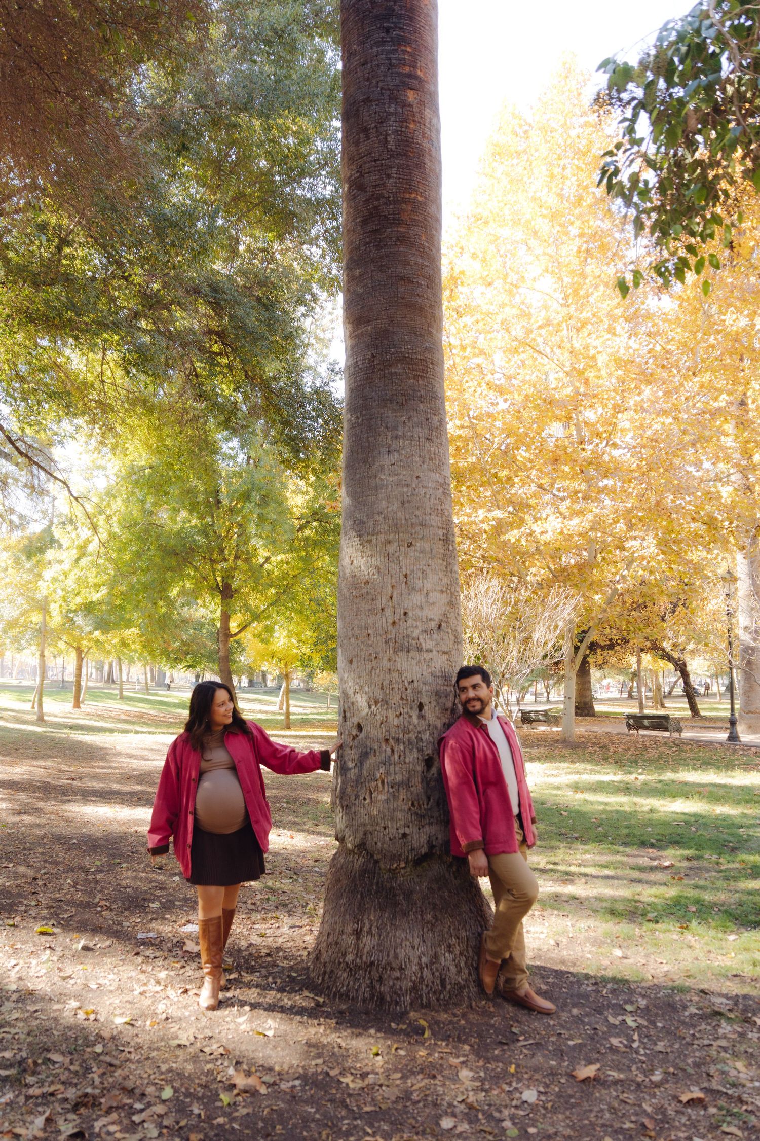 Pregnant couple in matching red jackets standing on opposite sides of a large tree in an autumn park.