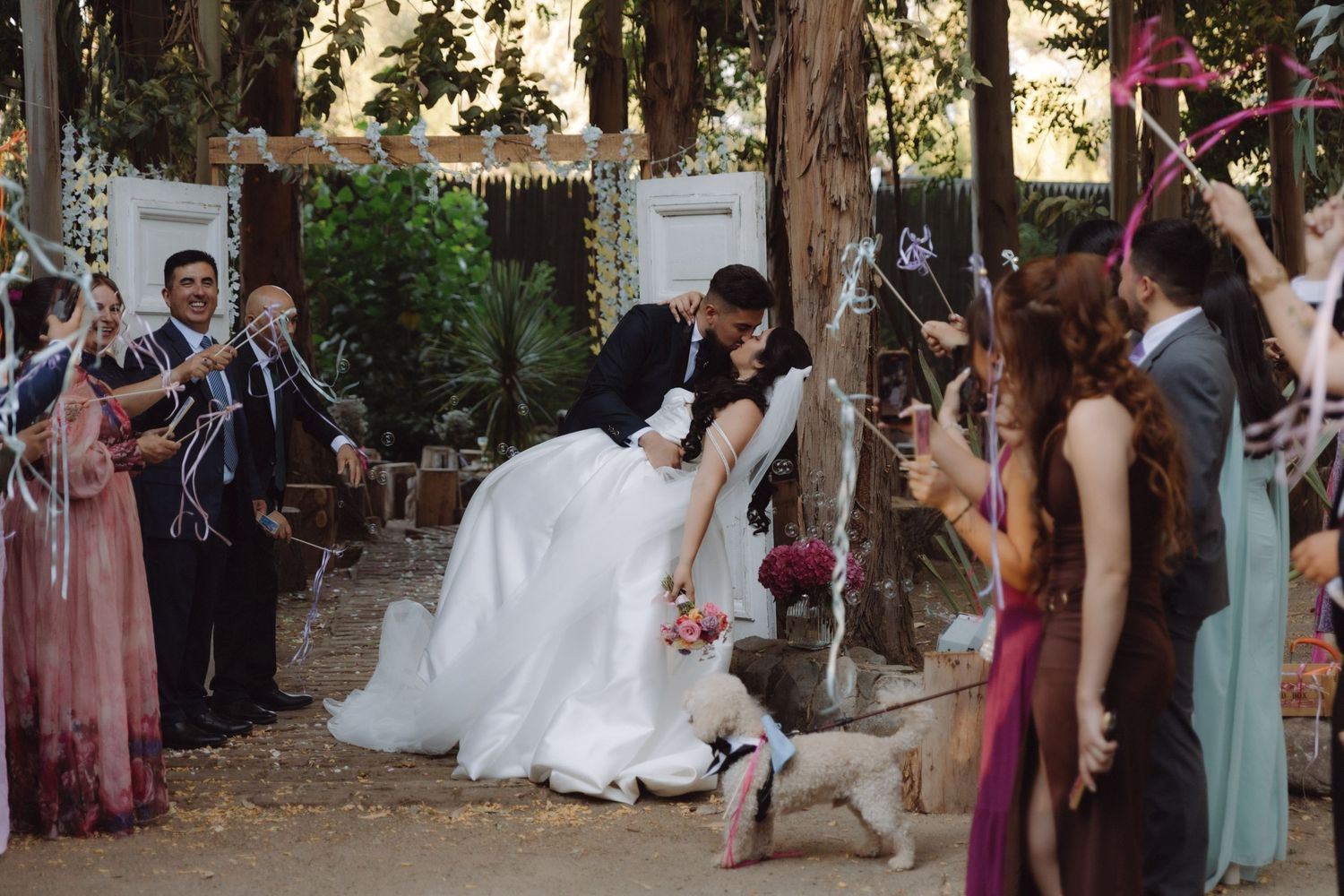 Bride and groom share a romantic dip kiss while guests celebrate with ribbons at rustic outdoor wedding ceremony.