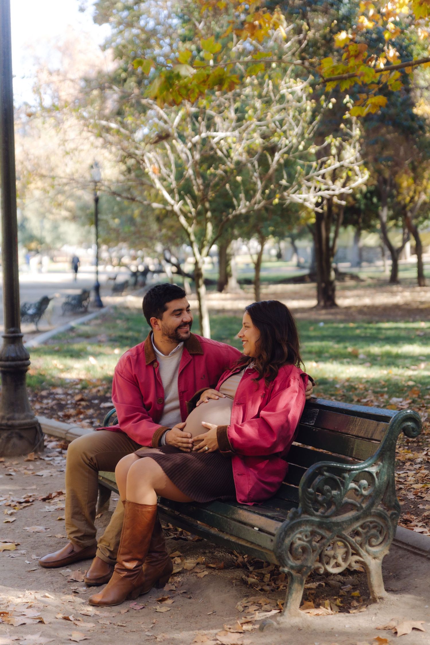 Smiling couple on a park bench in autumn, man tenderly touching pregnant woman's belly in warm sunlight.