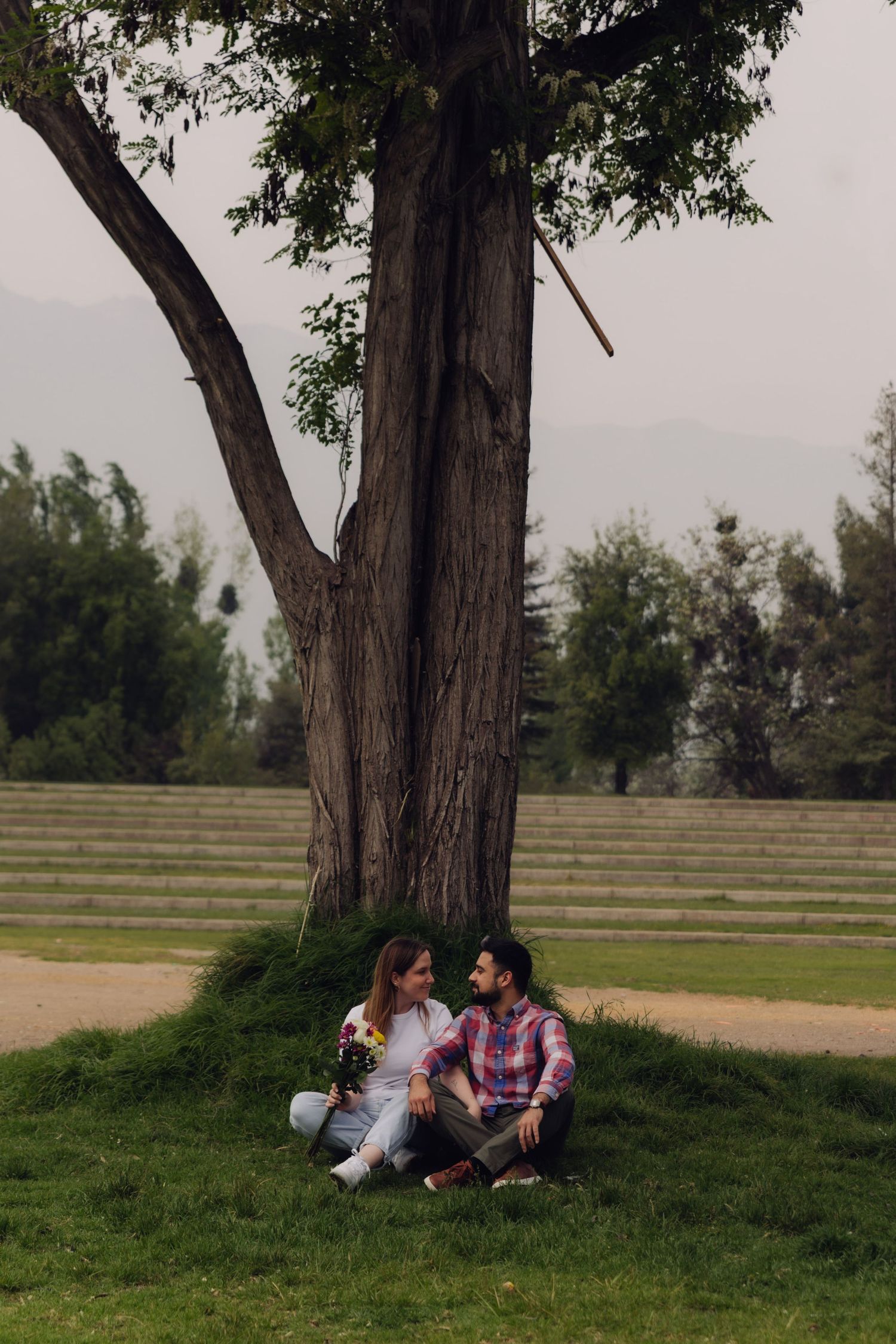 Romantic couple sitting together at the base of a large tree in a peaceful green park.