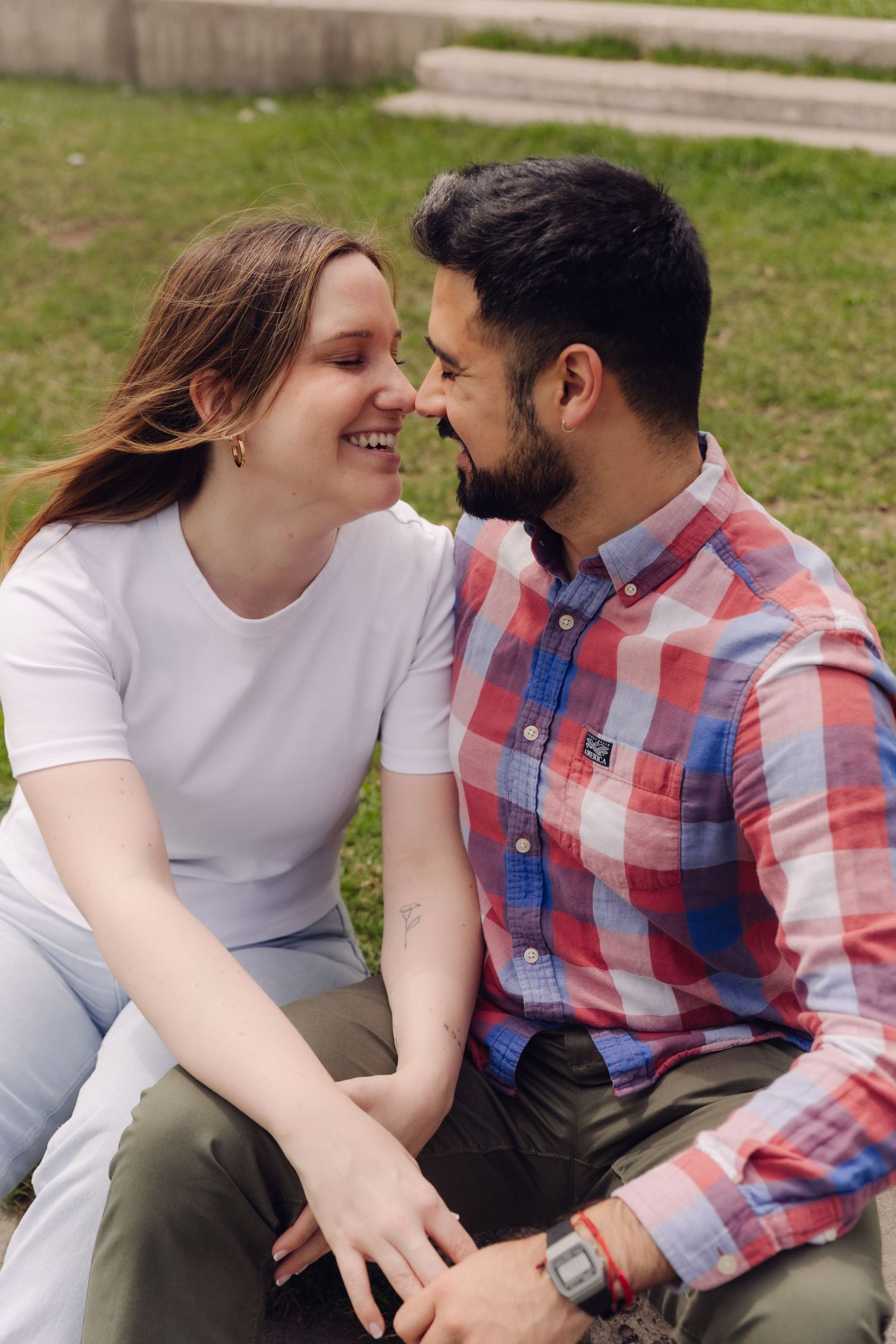 Smiling couple sitting on grass in a park, leaning close together in an intimate moment.