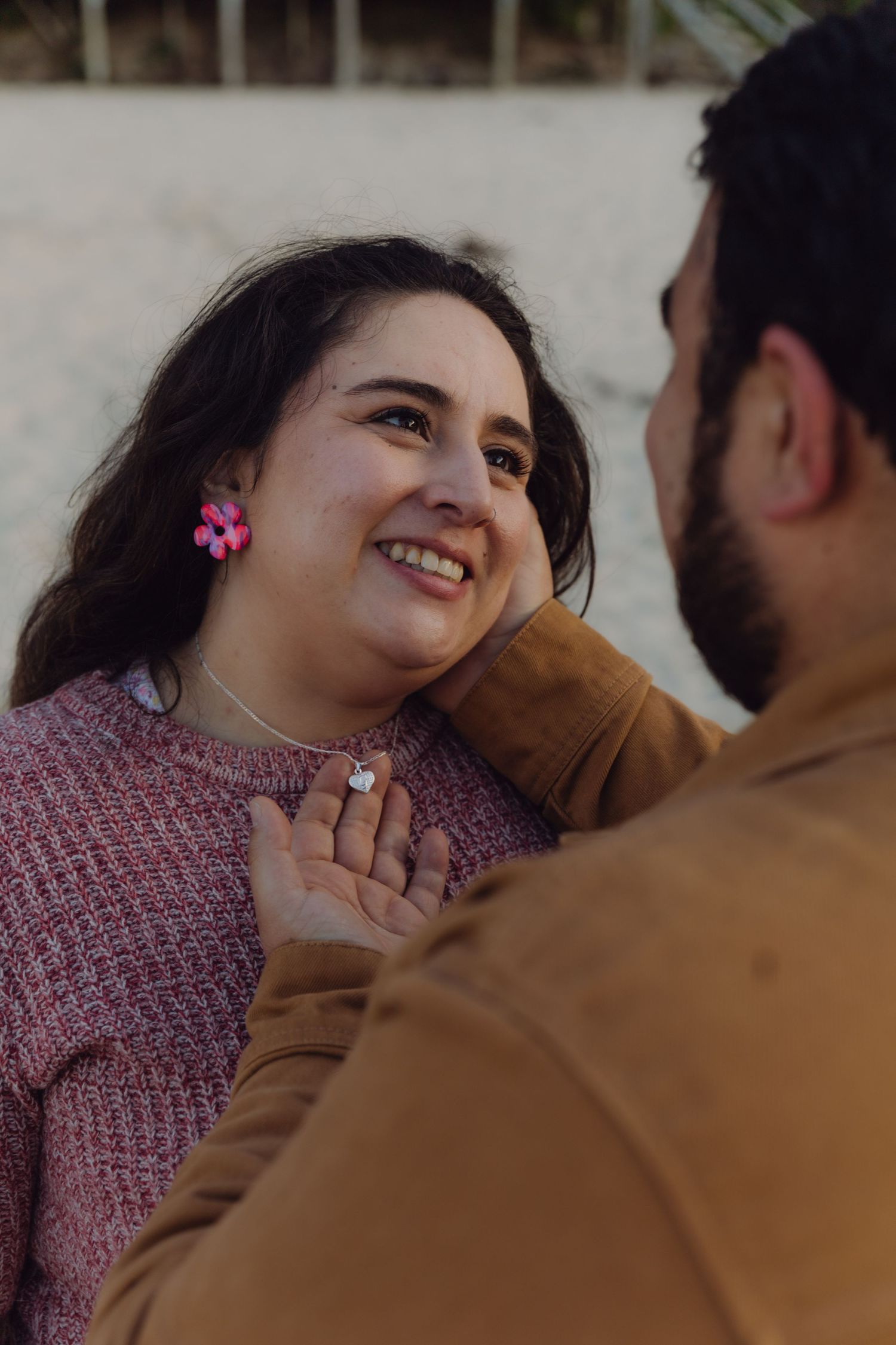 Man tenderly cupping smiling woman's face outdoors, with pink earrings and necklace visible.