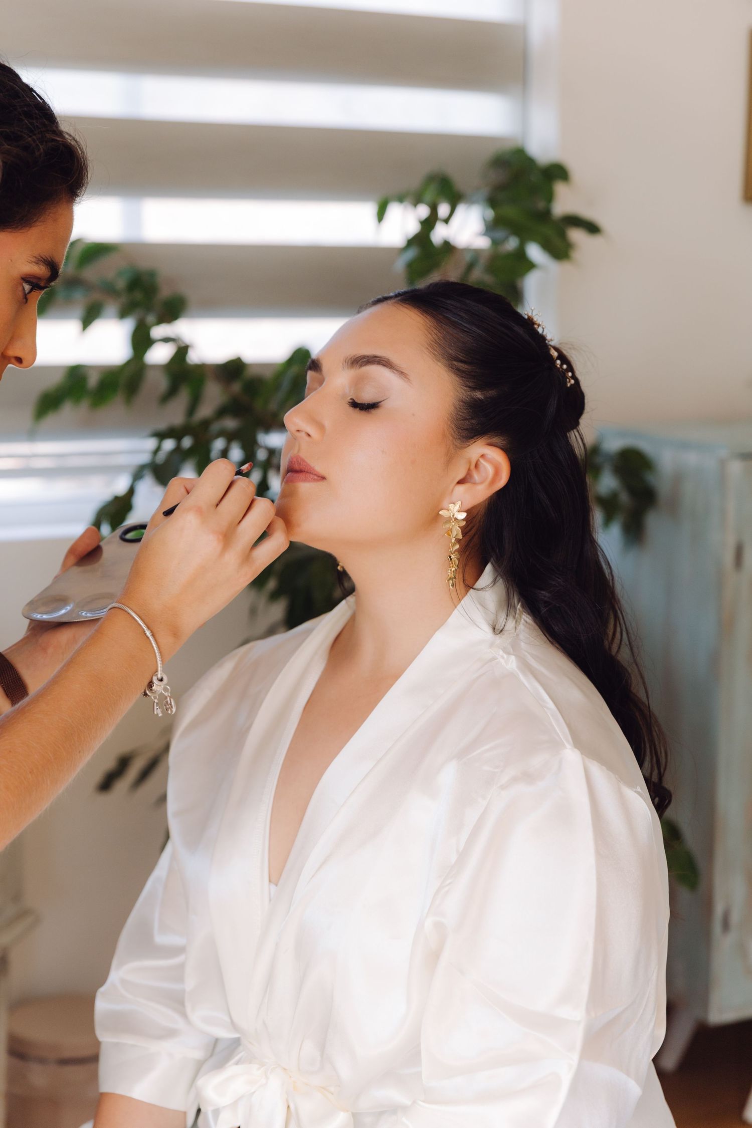 Makeup artist applying finishing touches to beautiful dark-haired bride in white satin robe.