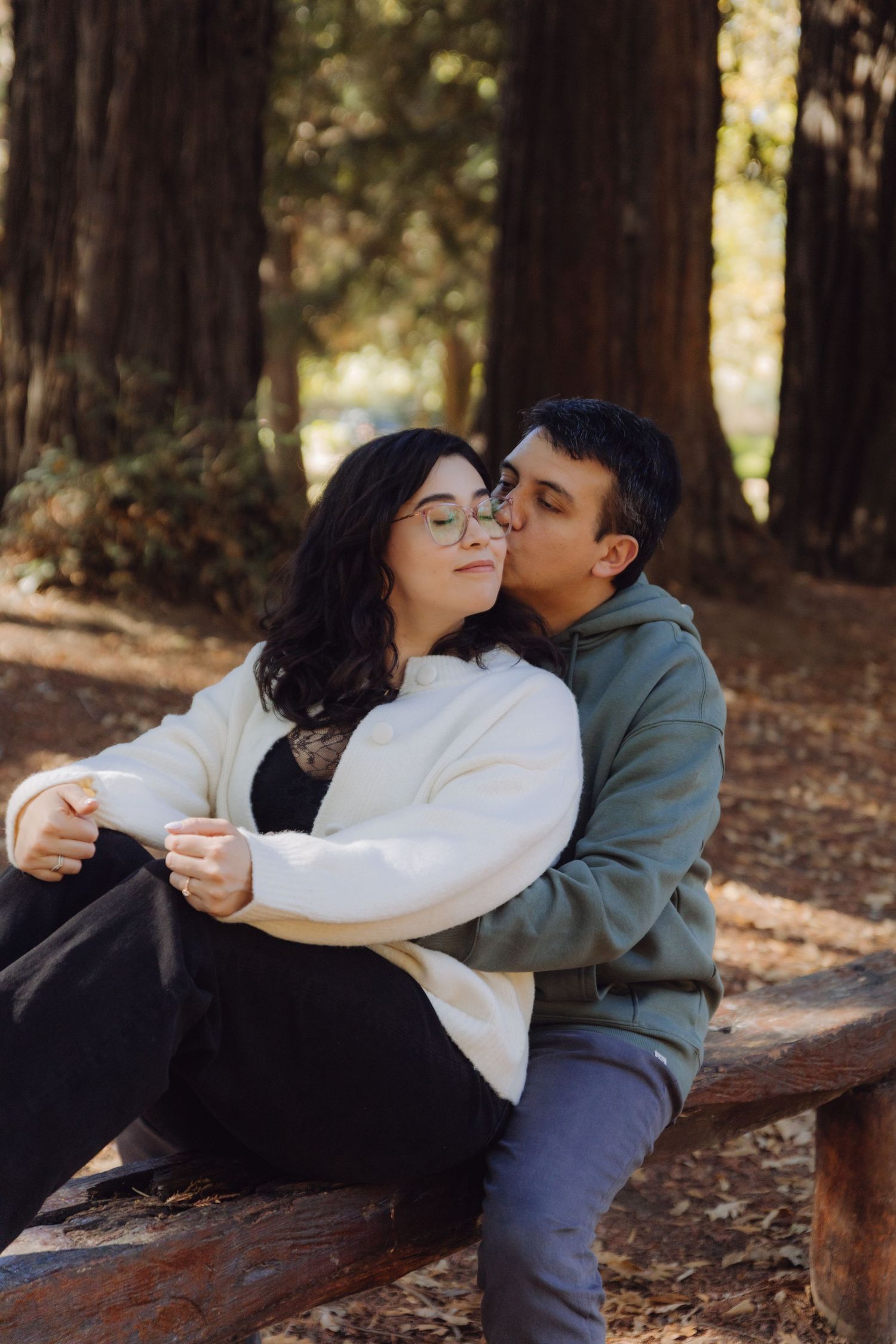 Couple sitting on a wooden bench in a redwood forest, sharing a tender kiss.