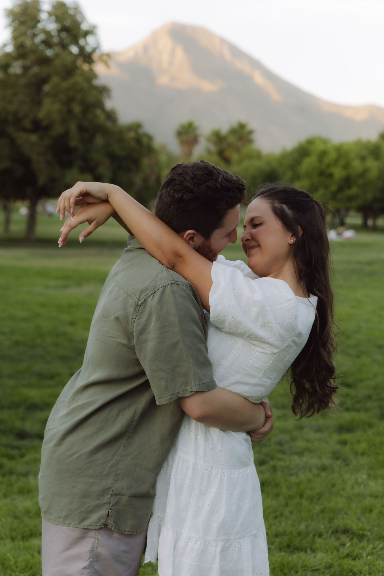 Couple embracing and almost kissing in green field with mountain backdrop at golden hour.