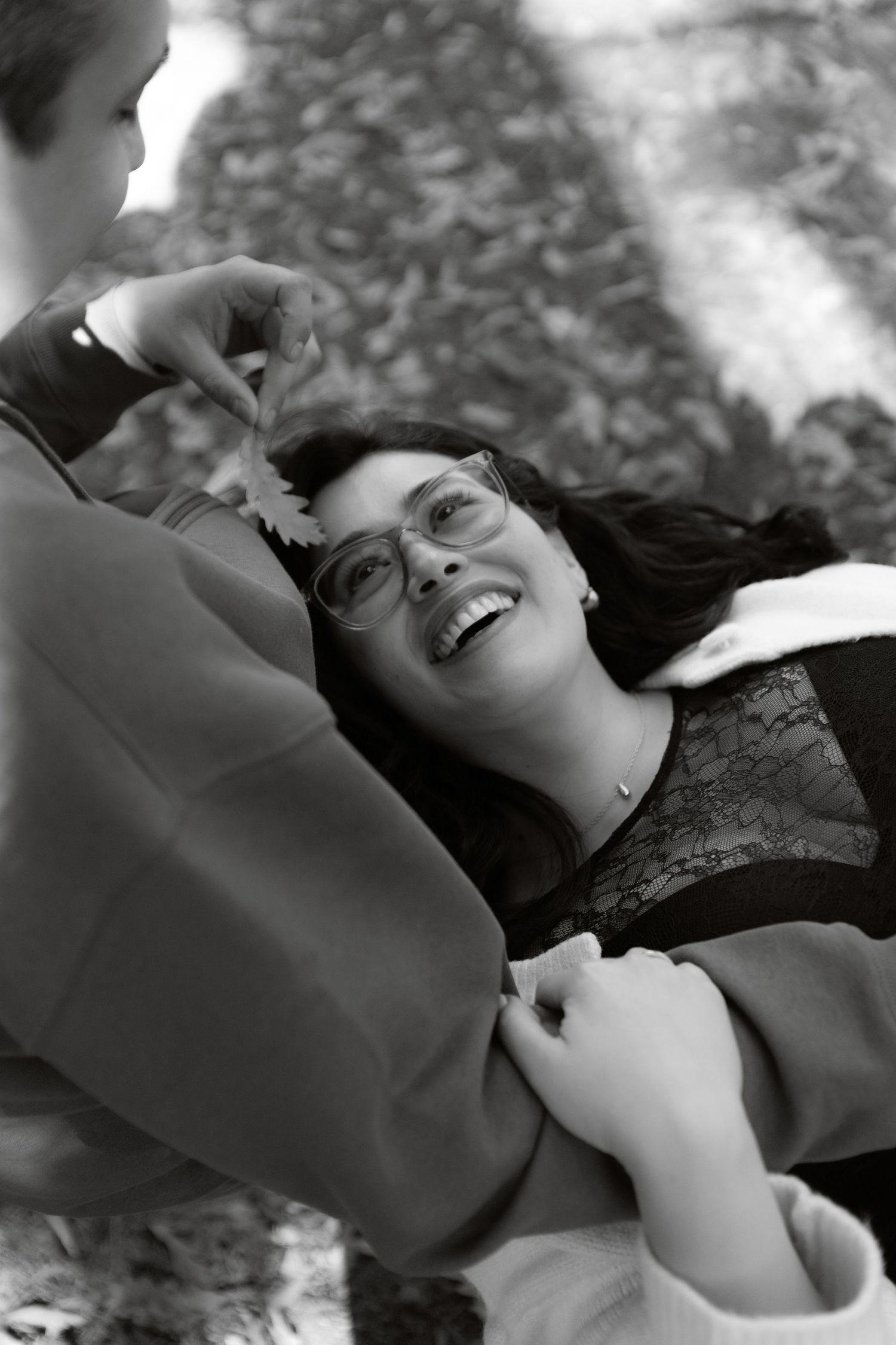 Black and white photo of a laughing woman with glasses lying back while partner holds a flower above her.