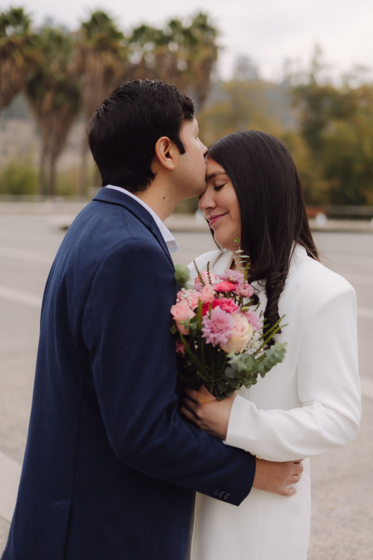 Groom in navy suit kissing bride on temple while she holds pink flower bouquet on urban street.