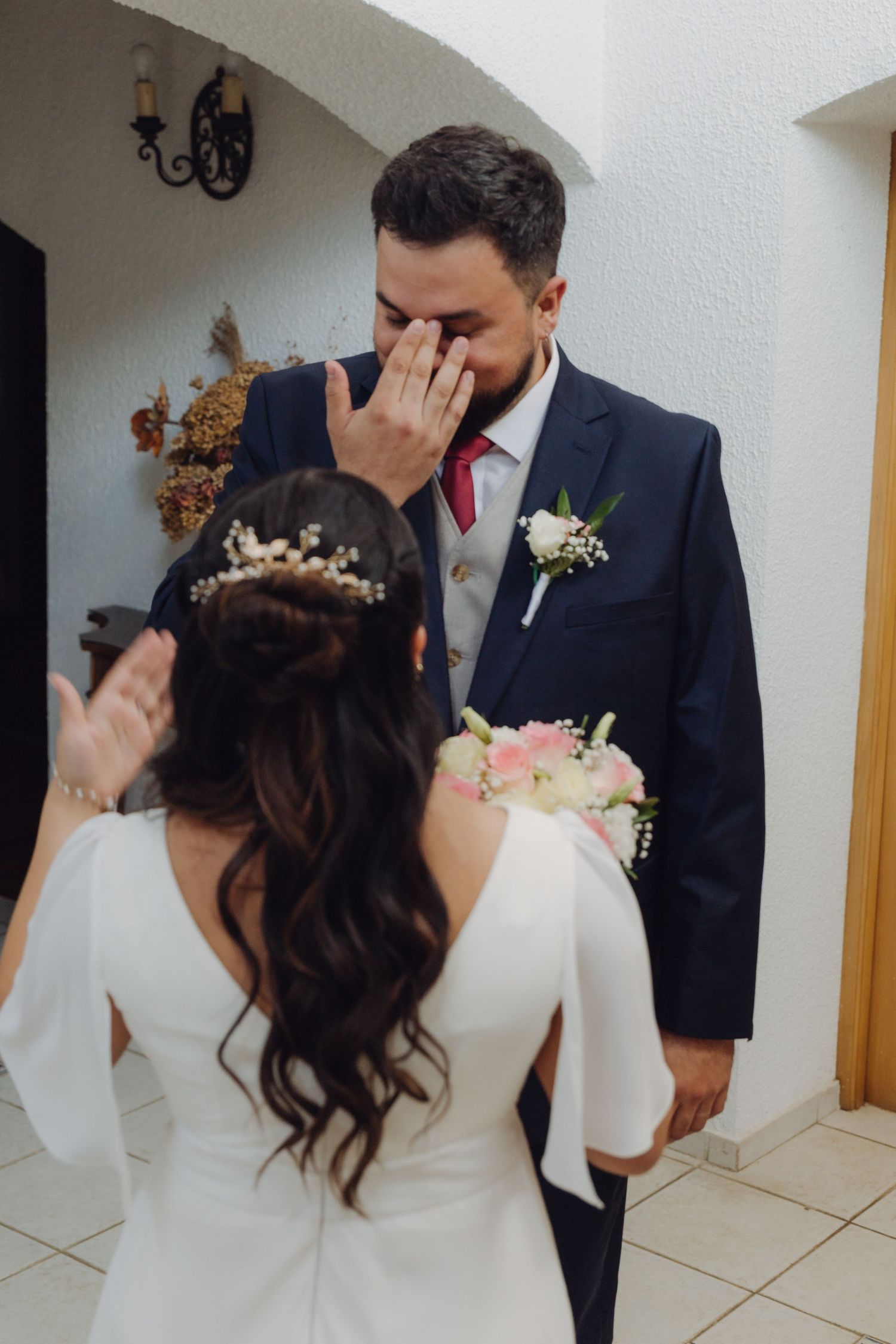 Emotional groom wiping tears as bride presents herself in white wedding dress during first look moment.