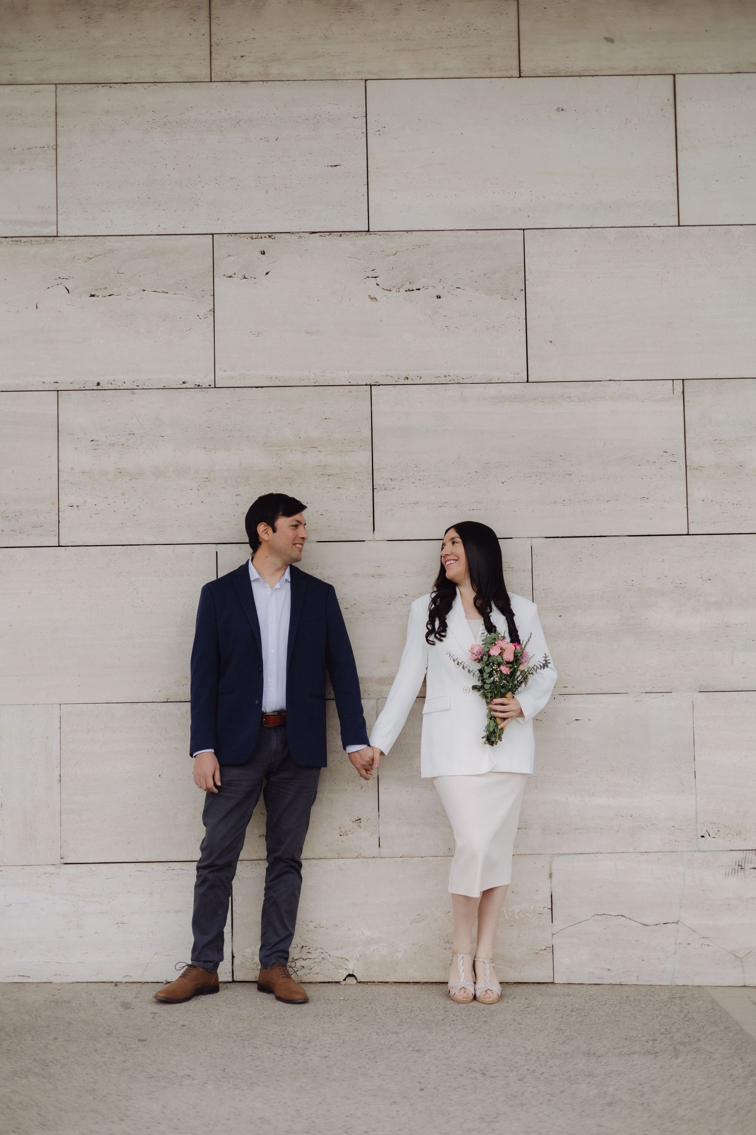 Couple holding hands in front of a stone wall, woman in white suit holding pink bouquet, man in navy blazer.