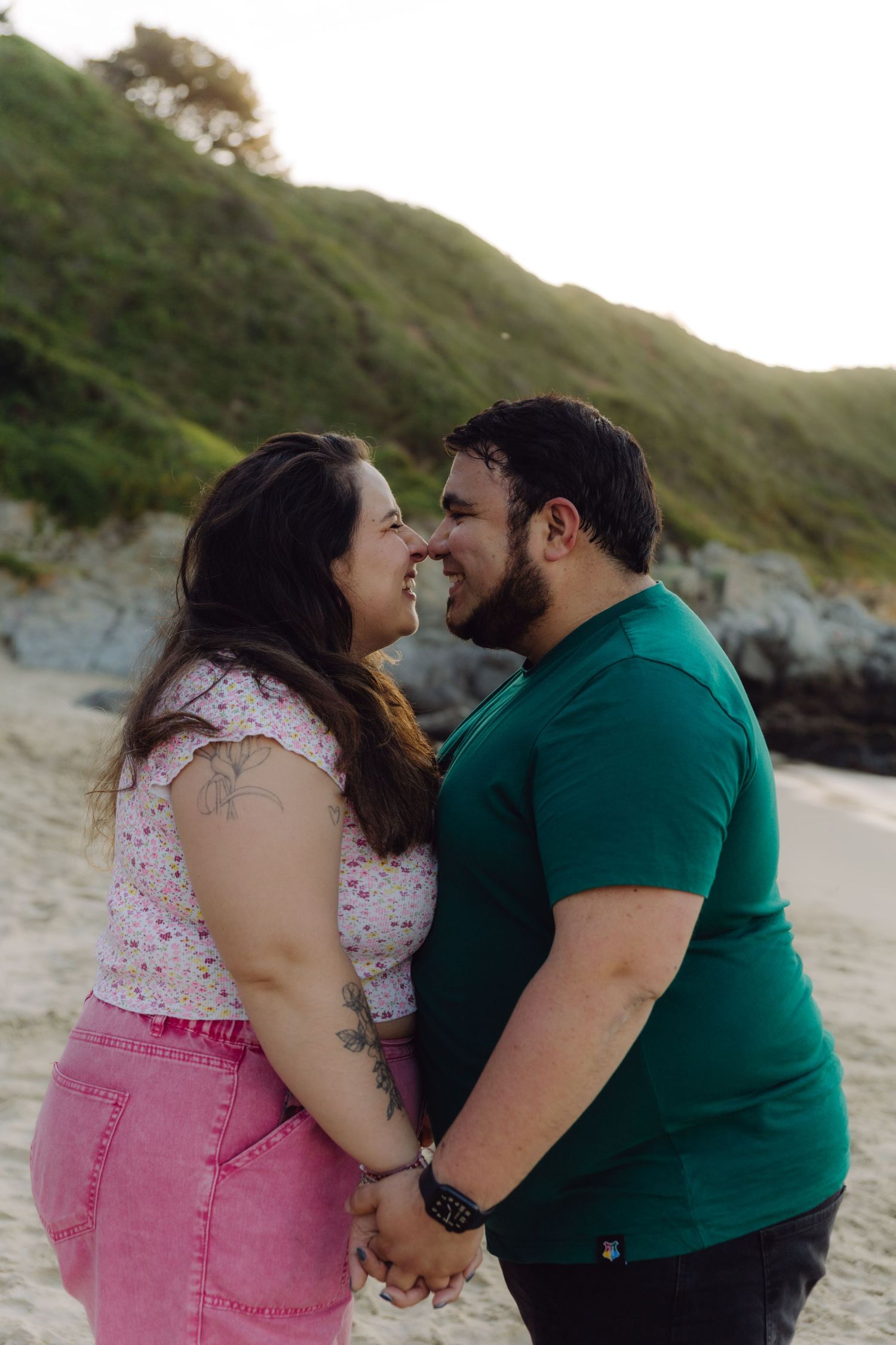 Happy couple holding hands and smiling at each other on a sandy beach with green hills in the background.