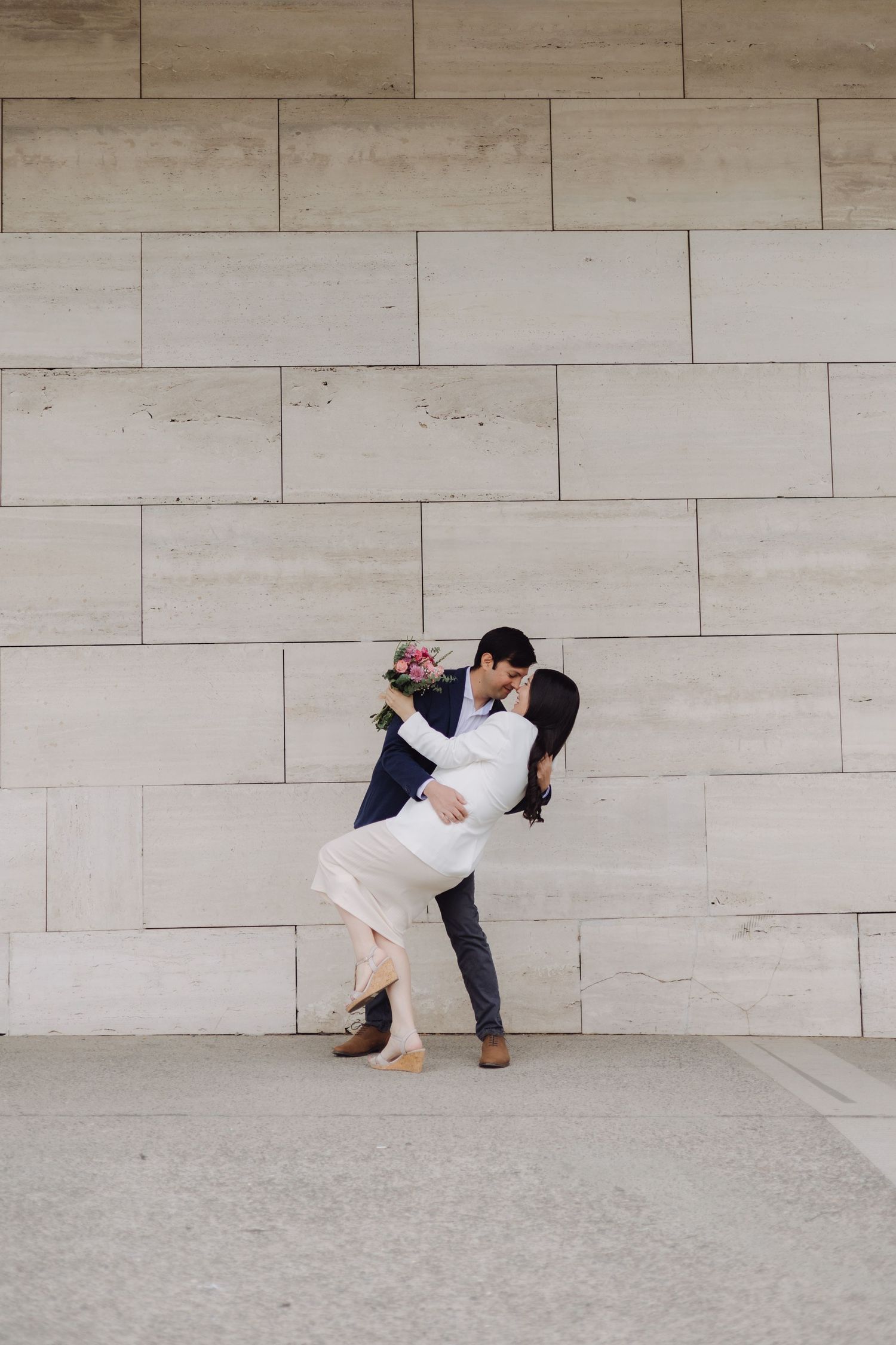Couple sharing a romantic dip kiss against a light stone wall, woman in white dress holding a floral bouquet.