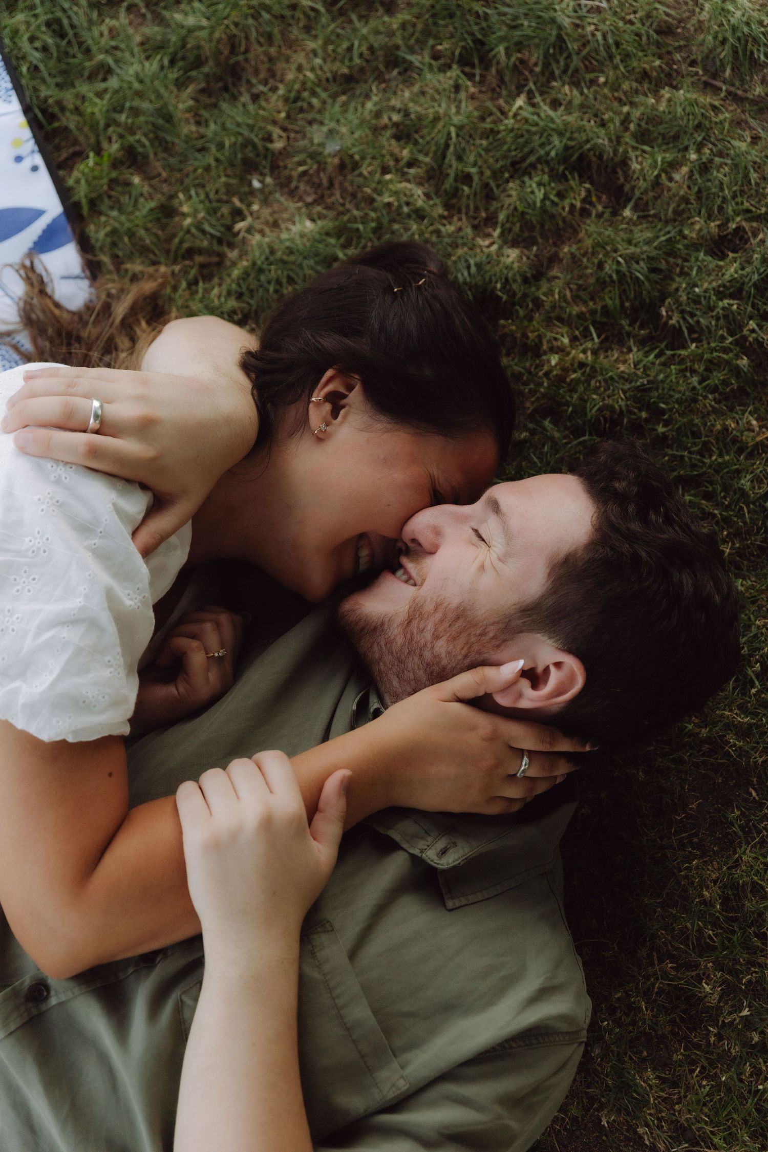 Couple lying on grass, sharing a kiss, woman in white dress with engagement ring visible.