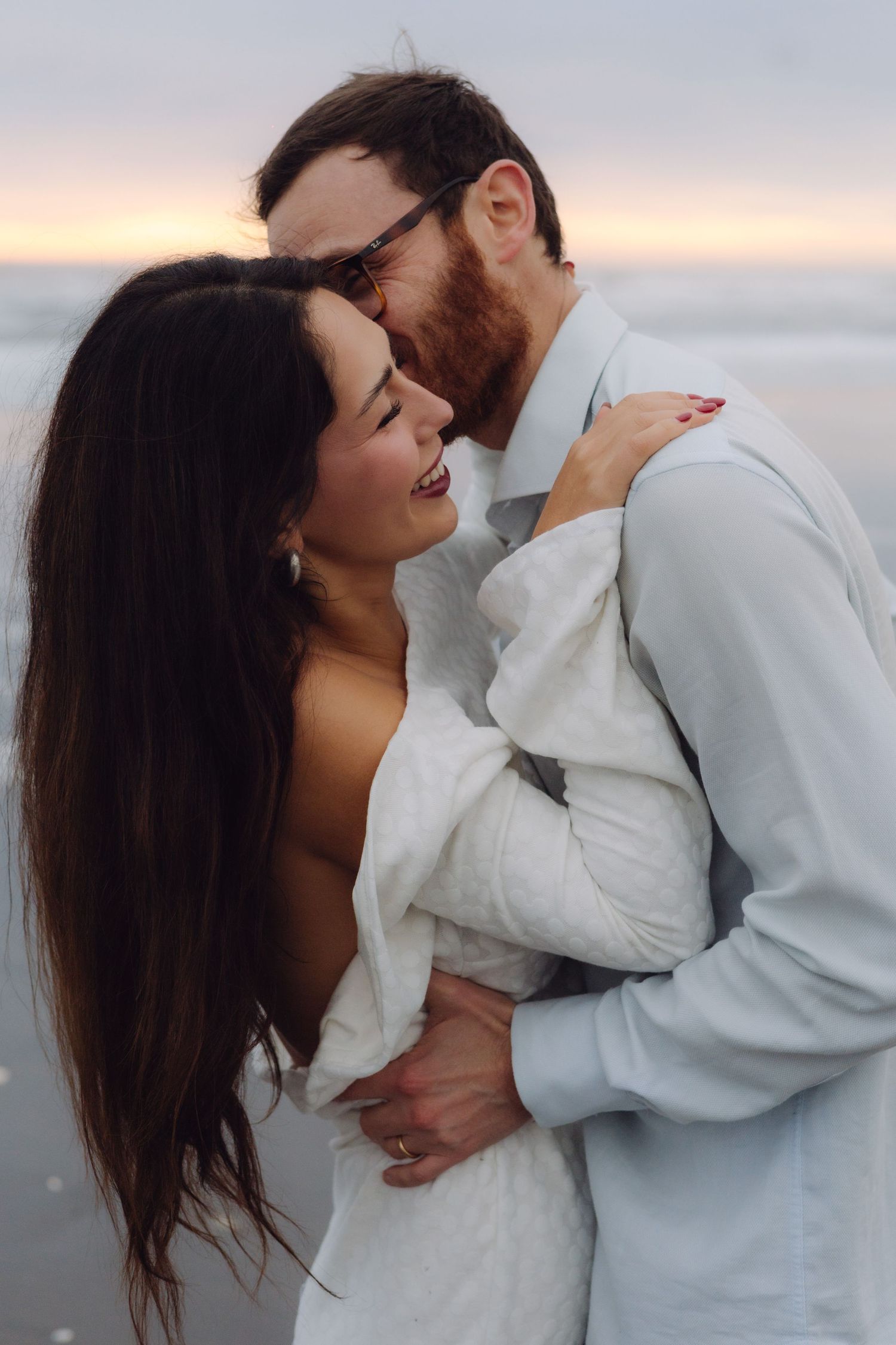Romantic couple embracing on a beach at dusk, man kissing woman's cheek as waves roll in behind them.