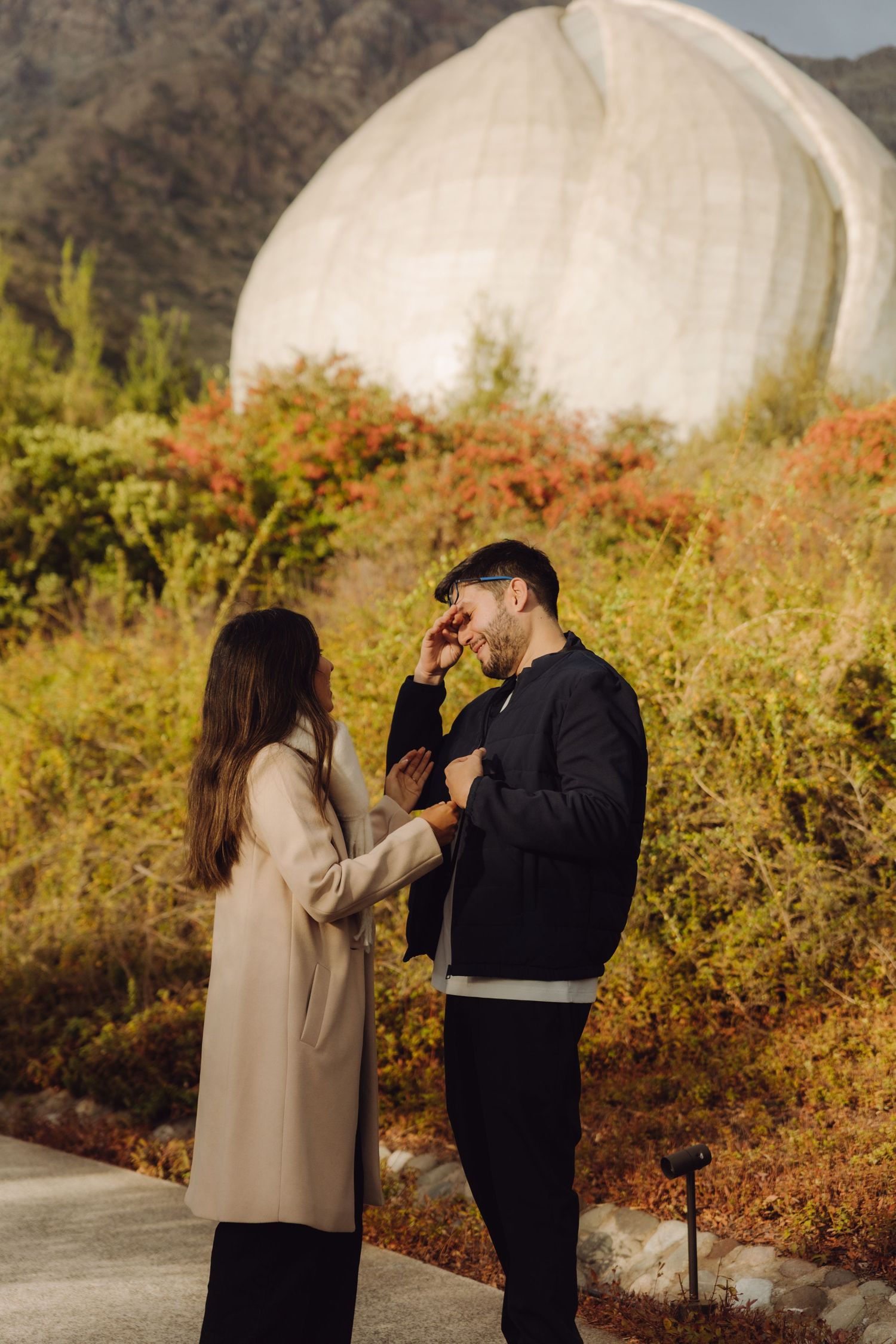 Couple sharing a tender moment outdoors with colorful autumn foliage and a white dome structure behind them.