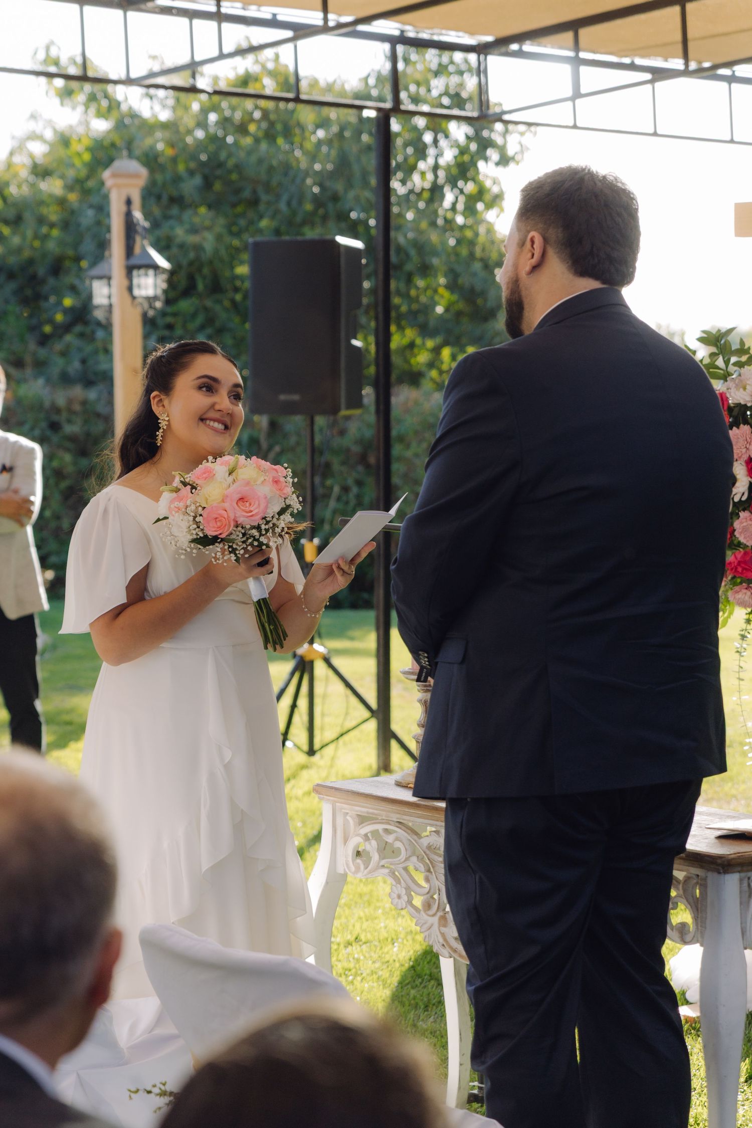 Smiling bride in white dress reads vows holding pink bouquet at outdoor wedding ceremony.