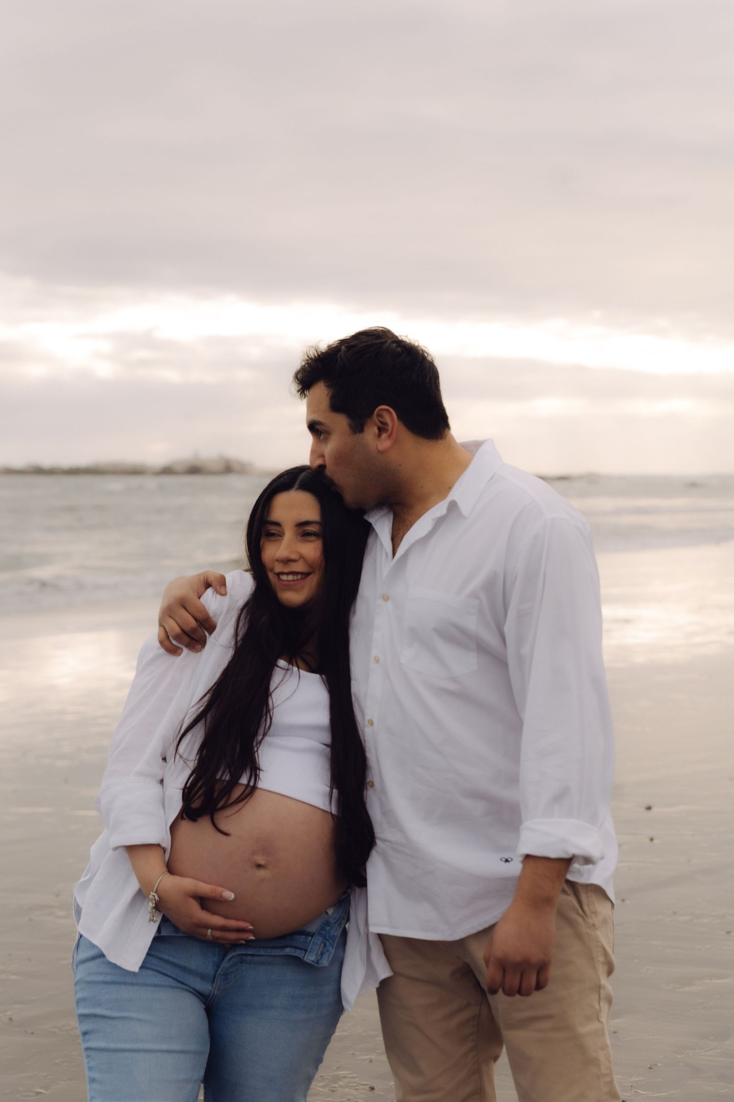 Pregnant couple embracing on beach at sunset, man kissing her head.