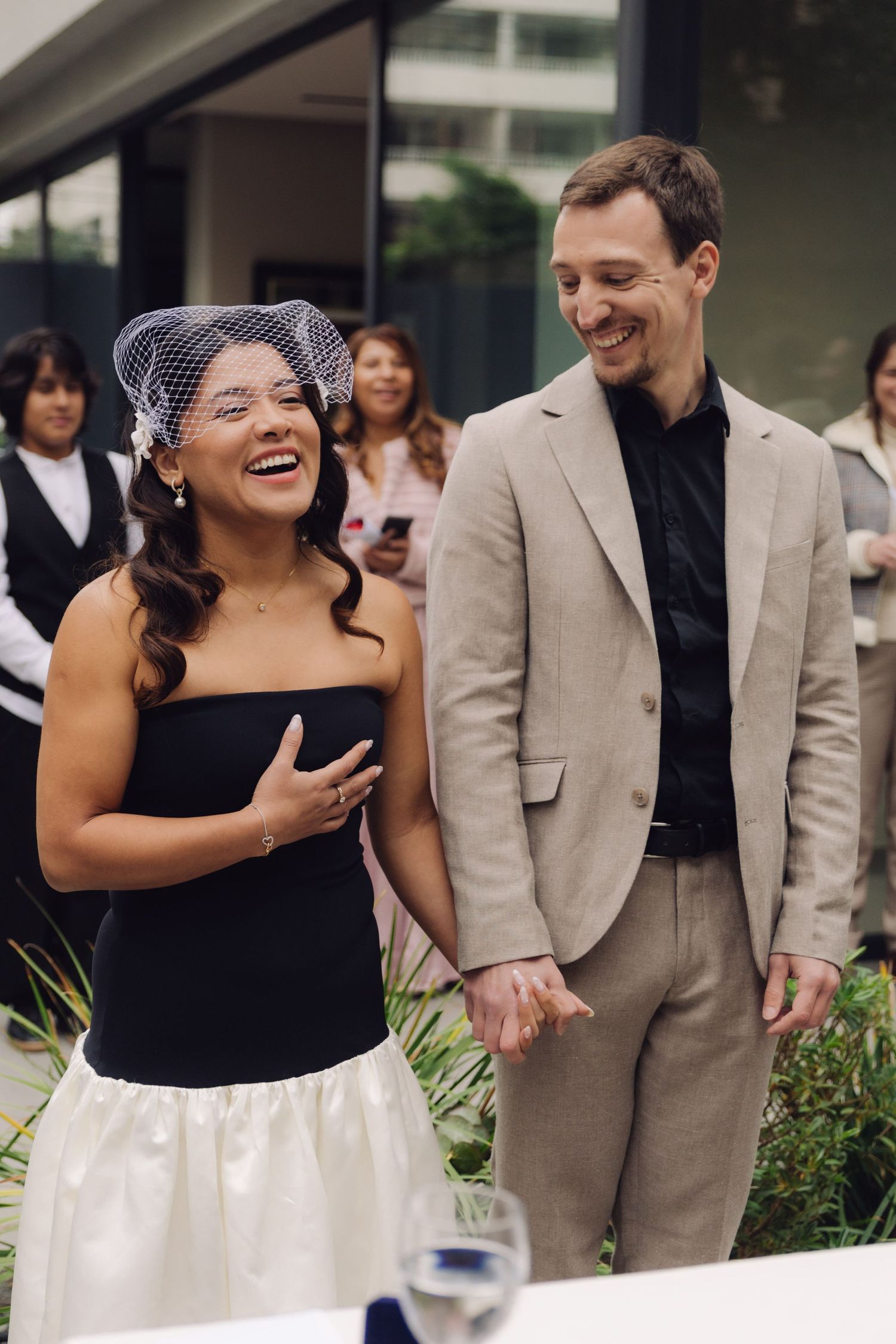 Joyful bride in black strapless dress with birdcage veil laughing with groom at outdoor wedding.