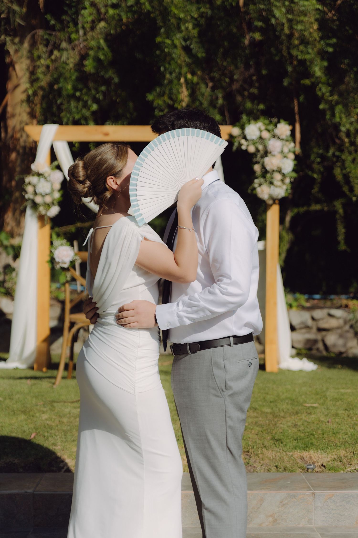 Bride and groom sharing a kiss hidden behind a white hand fan at outdoor wedding ceremony.