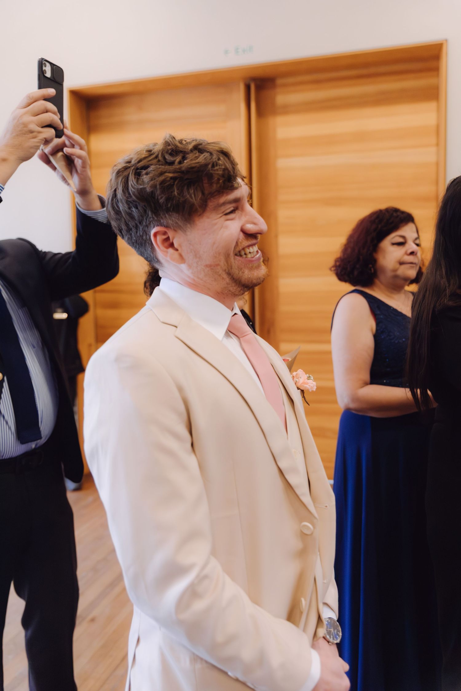 Smiling groom in cream suit with pink tie laughing joyfully at wedding ceremony indoors.
