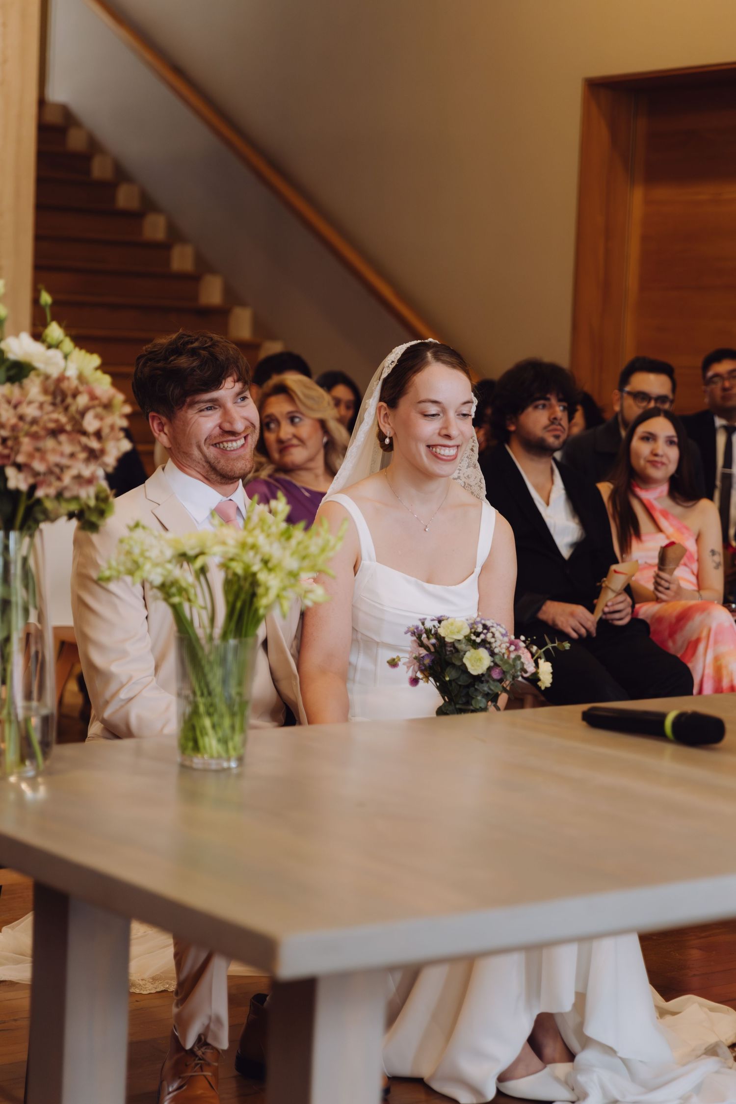 Happy bride and groom sitting together at wedding ceremony table, smiling with guests behind.