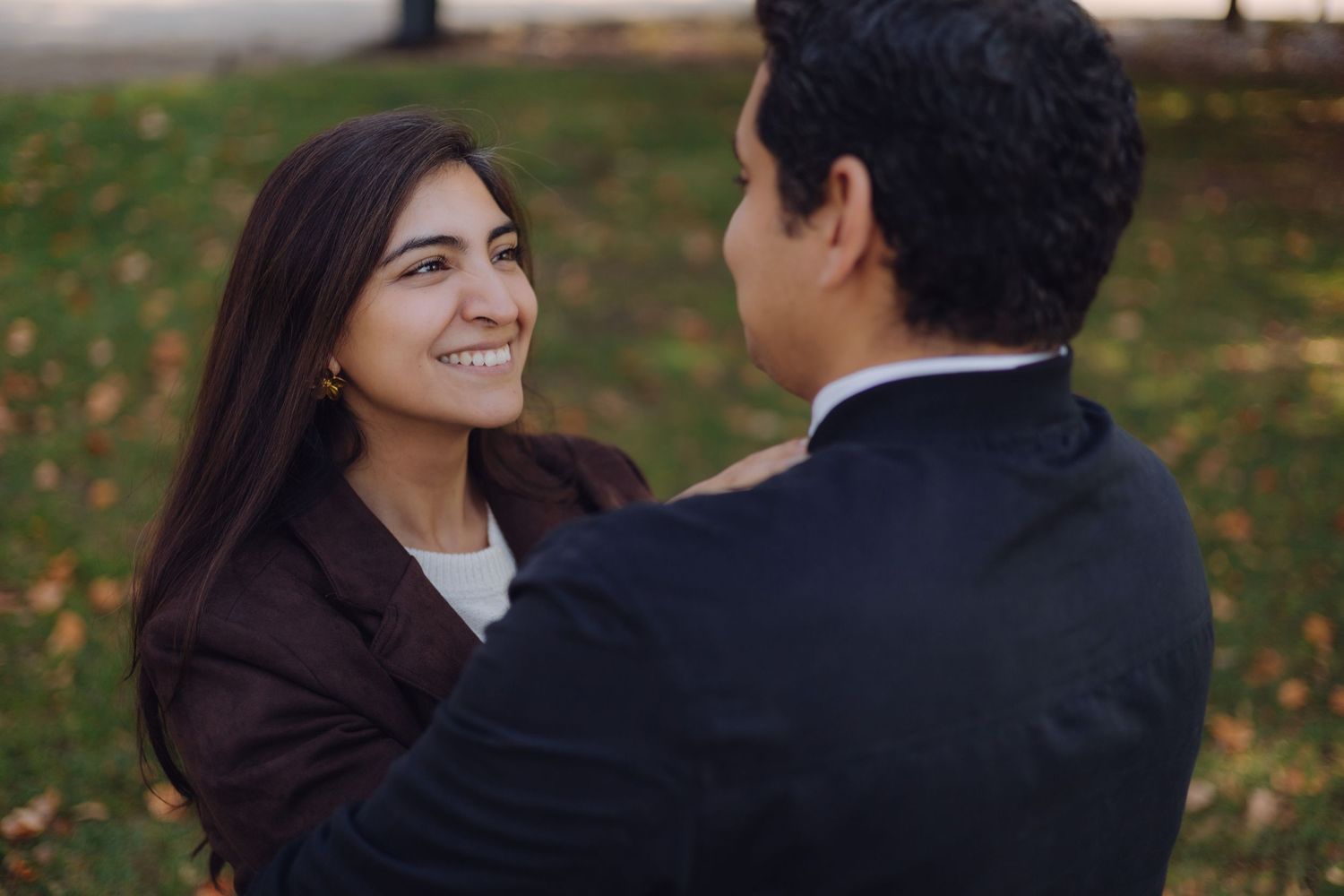 Smiling woman in brown jacket gazes at partner during romantic outdoor engagement session in autumn park.