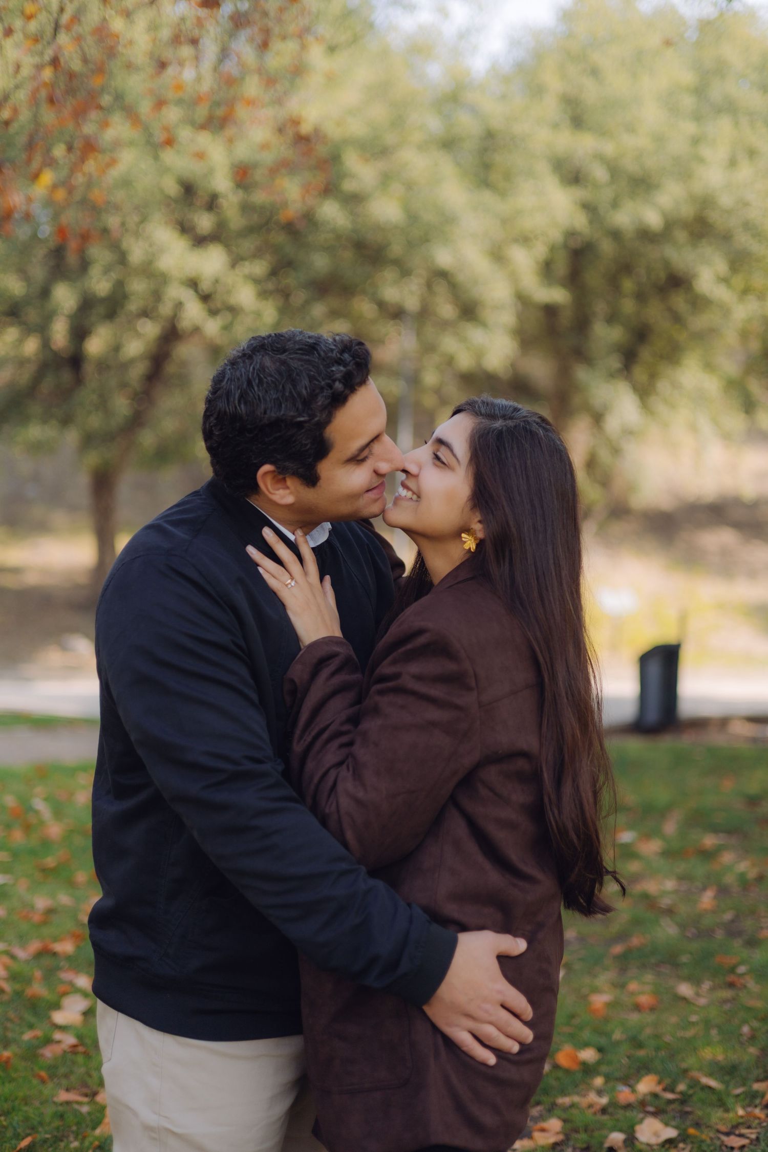 Couple sharing a romantic kiss in an autumn park, woman in brown jacket smiling with yellow earrings.