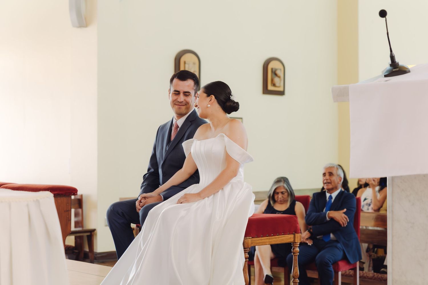 Bride and groom sitting together at church altar during wedding ceremony with nun watching.