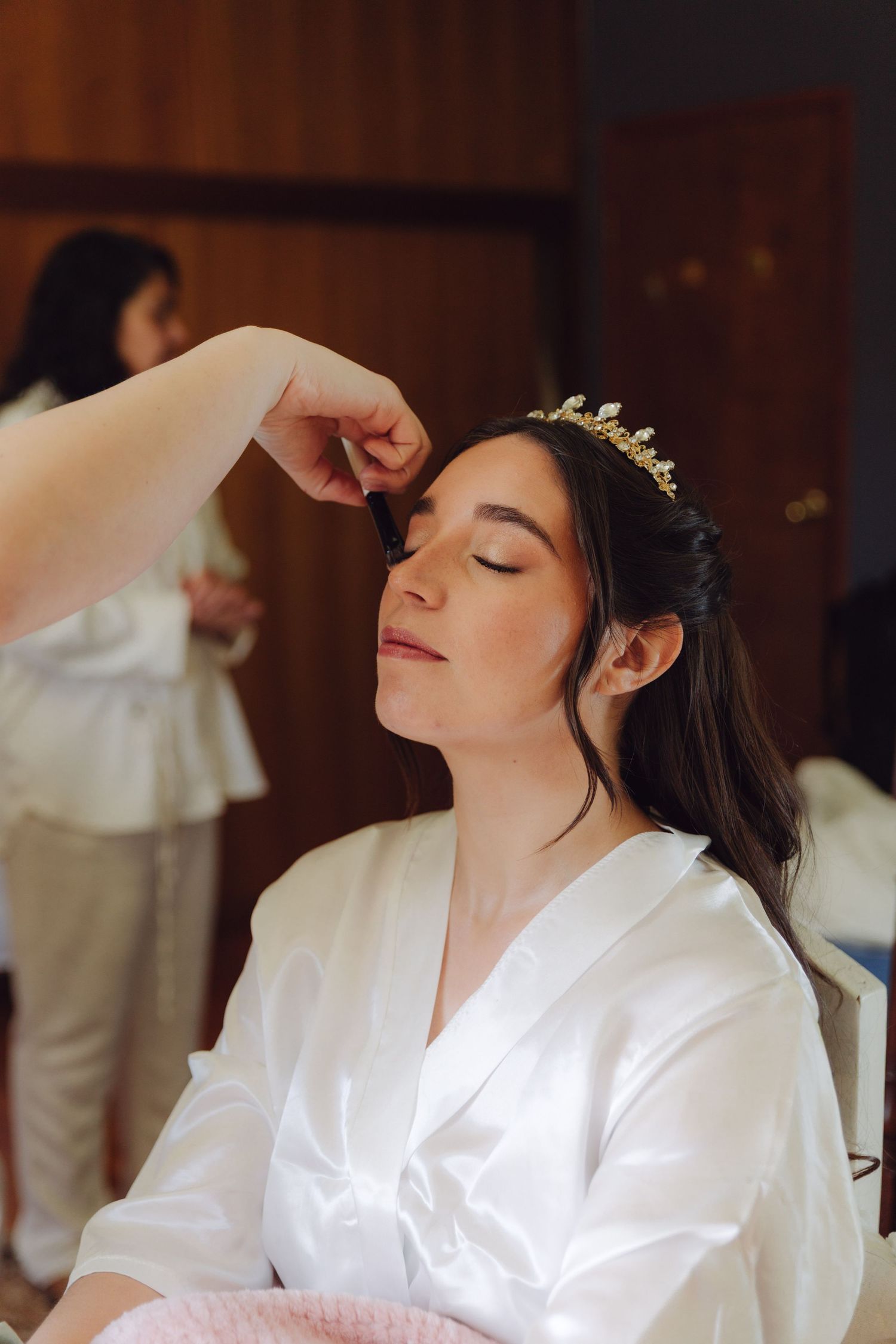 Bride getting makeup applied while wearing a white satin robe and gold tiara before her wedding ceremony.