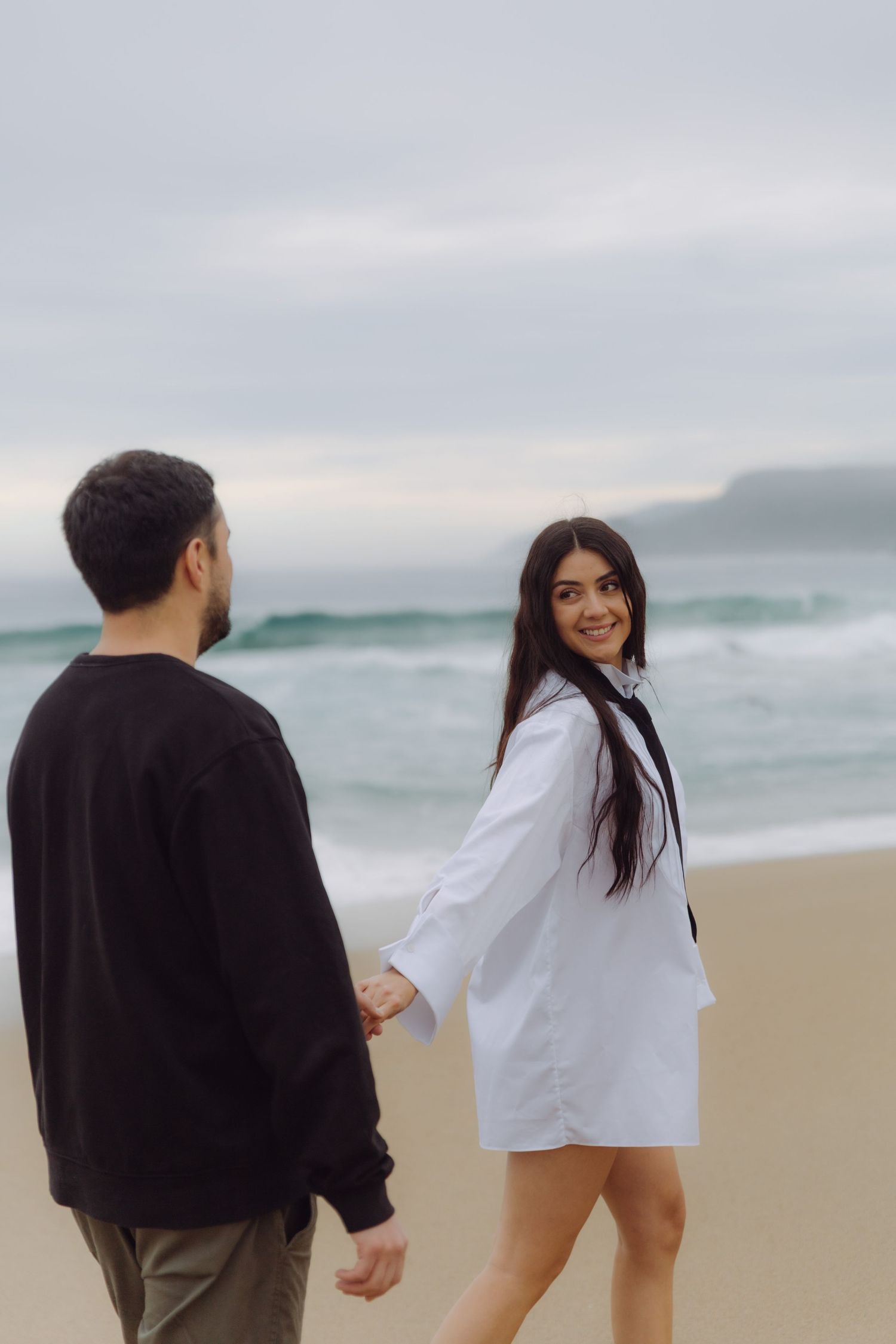 Smiling woman in white shirt looks back while holding hands with man walking on sandy beach.