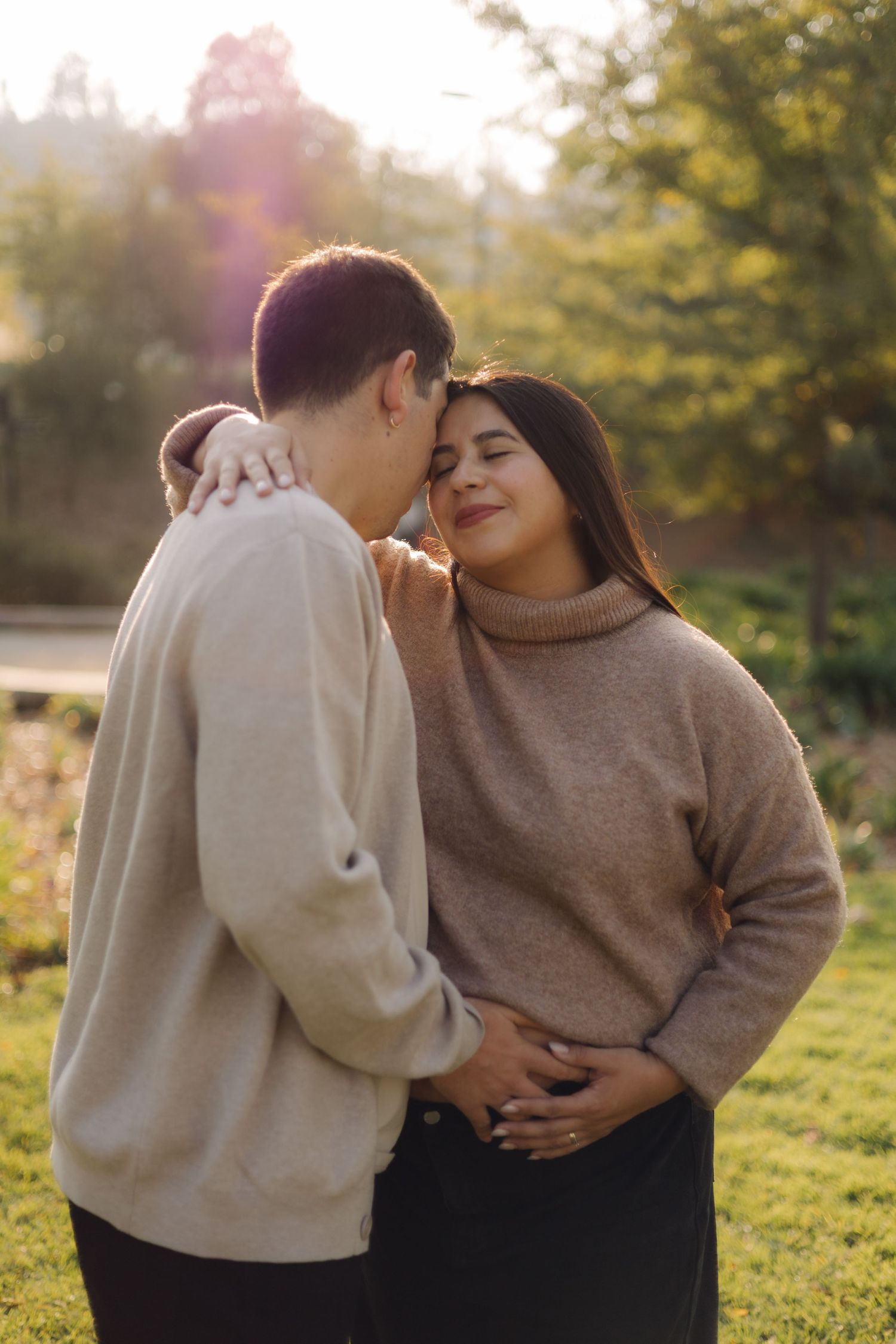 Couple sharing a tender kiss in a sunlit park, woman in beige turtleneck smiling with eyes closed.
