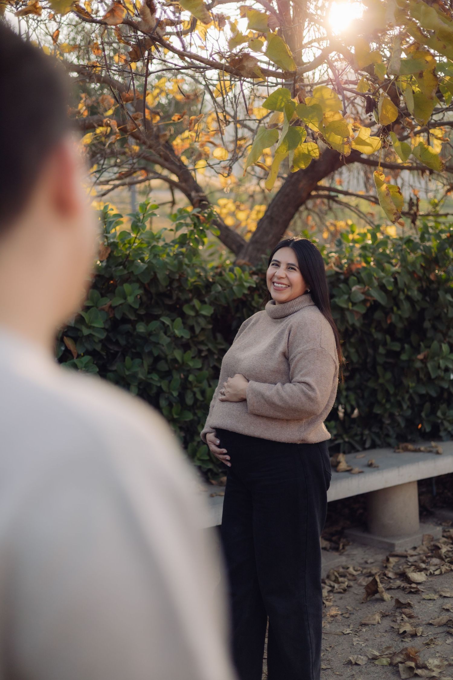 Smiling pregnant woman in beige sweater holds her belly while standing in a sunlit autumn park.