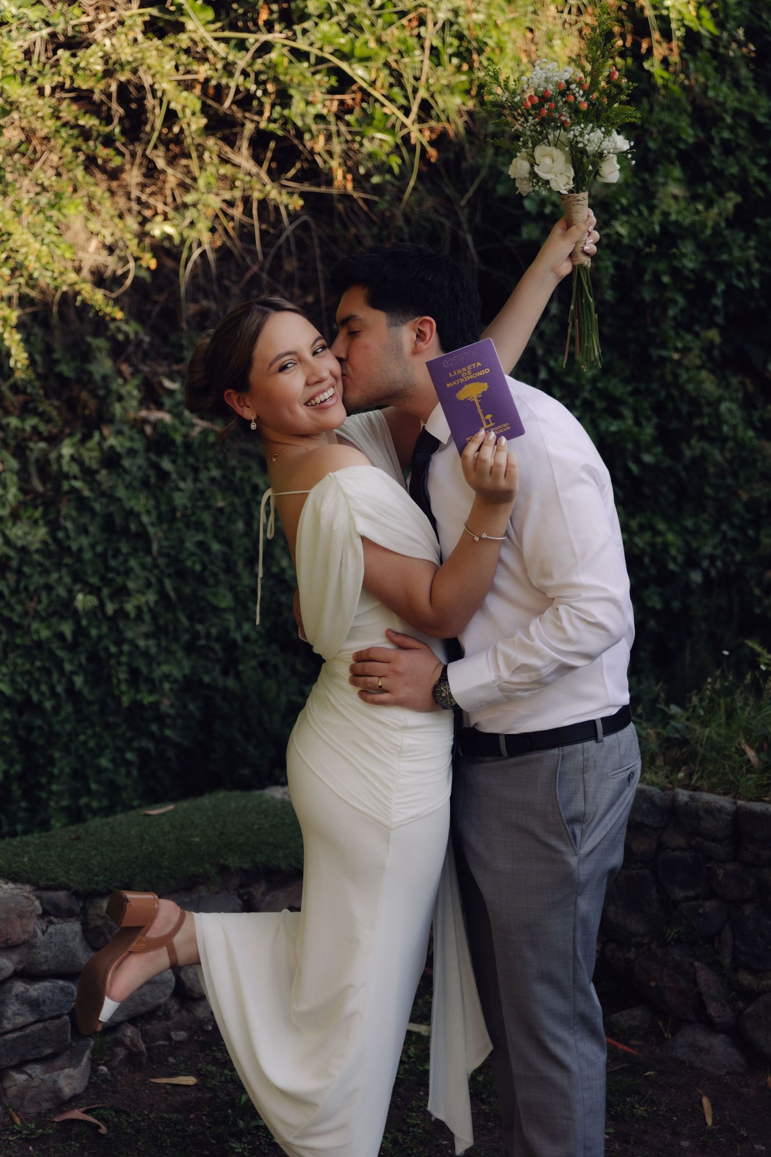 Smiling bride in white dress holding bouquet and marriage booklet while groom kisses her cheek.