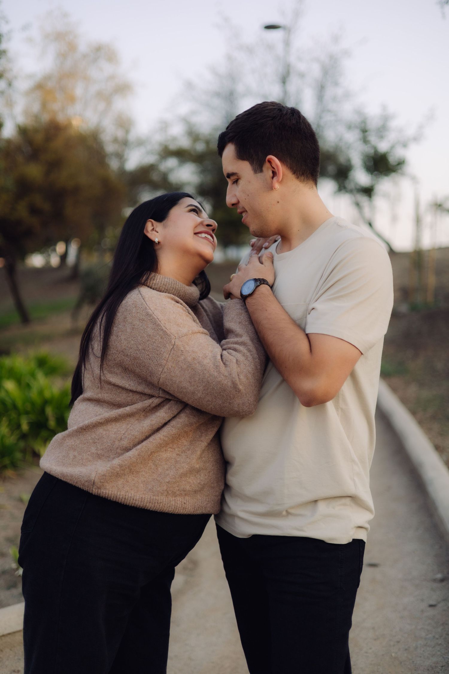 Happy couple holding hands and gazing at each other on a park pathway at dusk in casual sweaters.